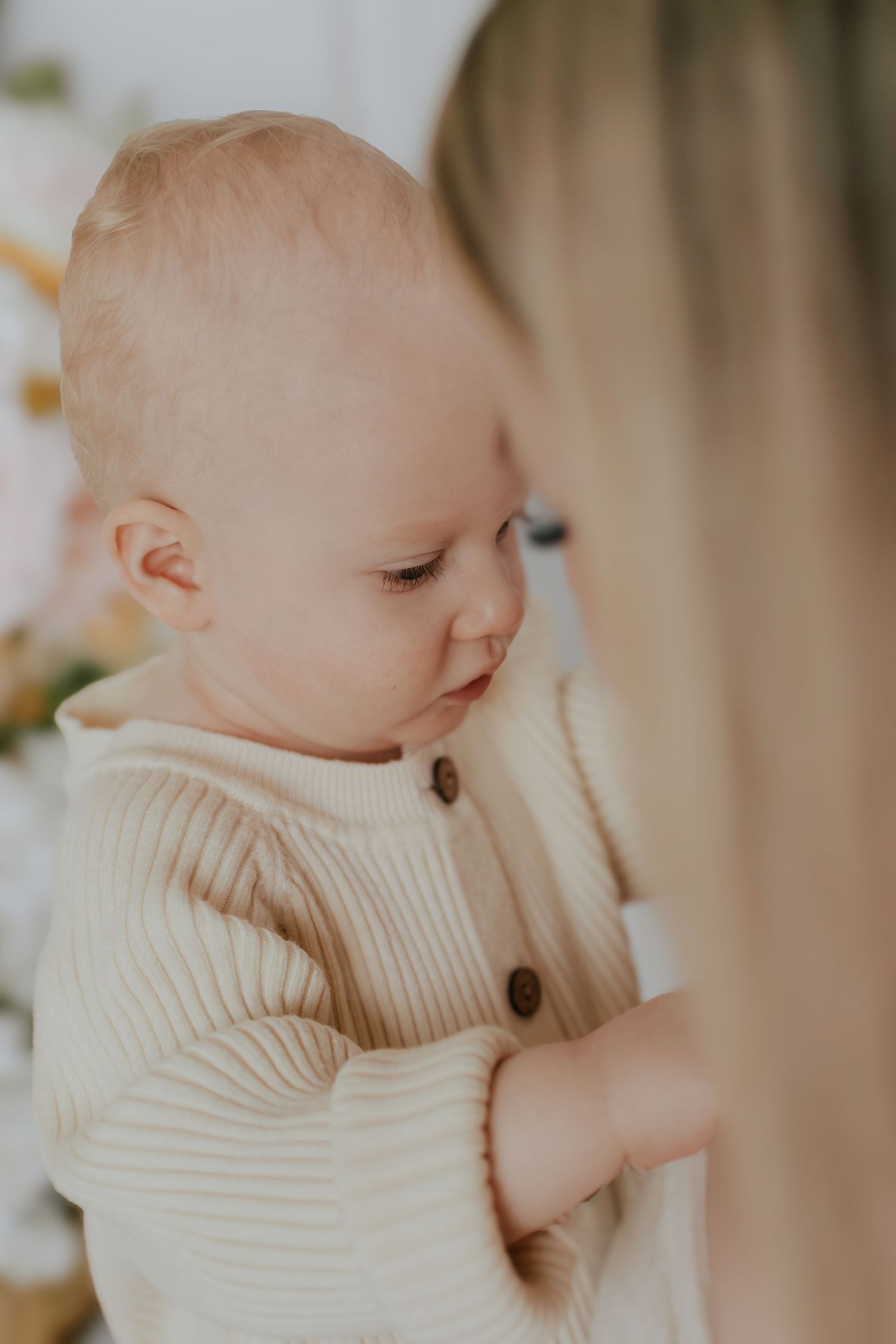 A blonde woman holds a cute baby dressed in a cream ribbed button-up outfit, both looking downward in soft light.