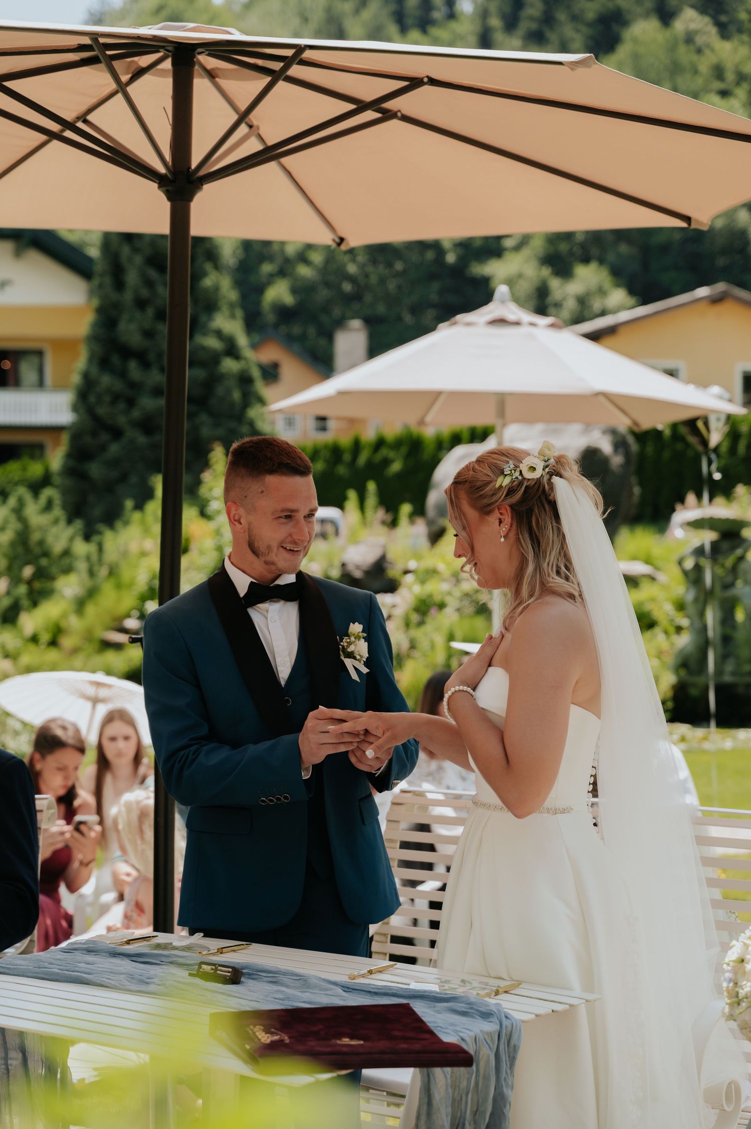 Groom in blue tuxedo placing wedding ring on brides finger during outdoor ceremony.