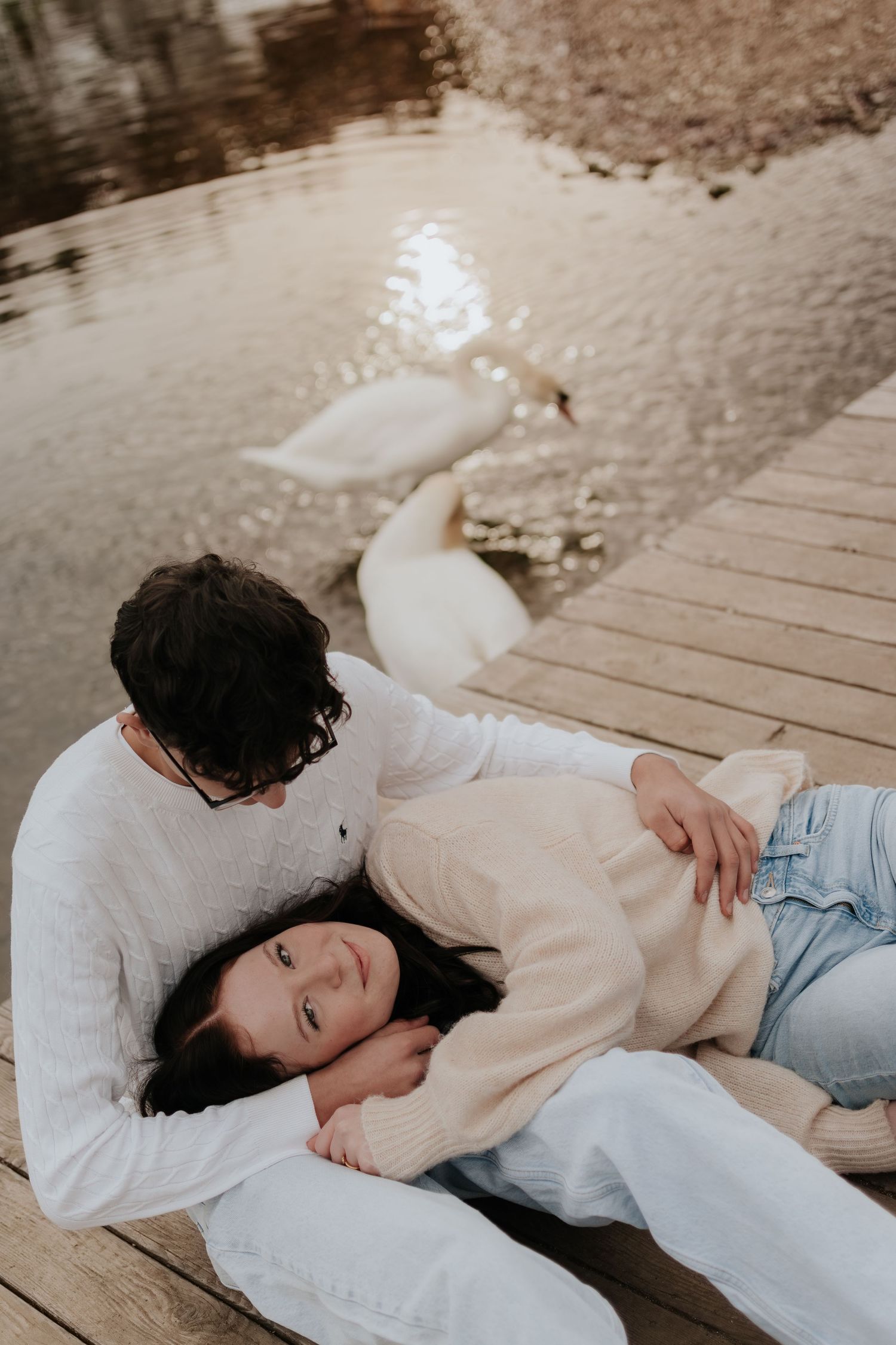 Couple relaxing on a wooden dock by a lake with swans, wearing cozy sweaters in a romantic outdoor setting.