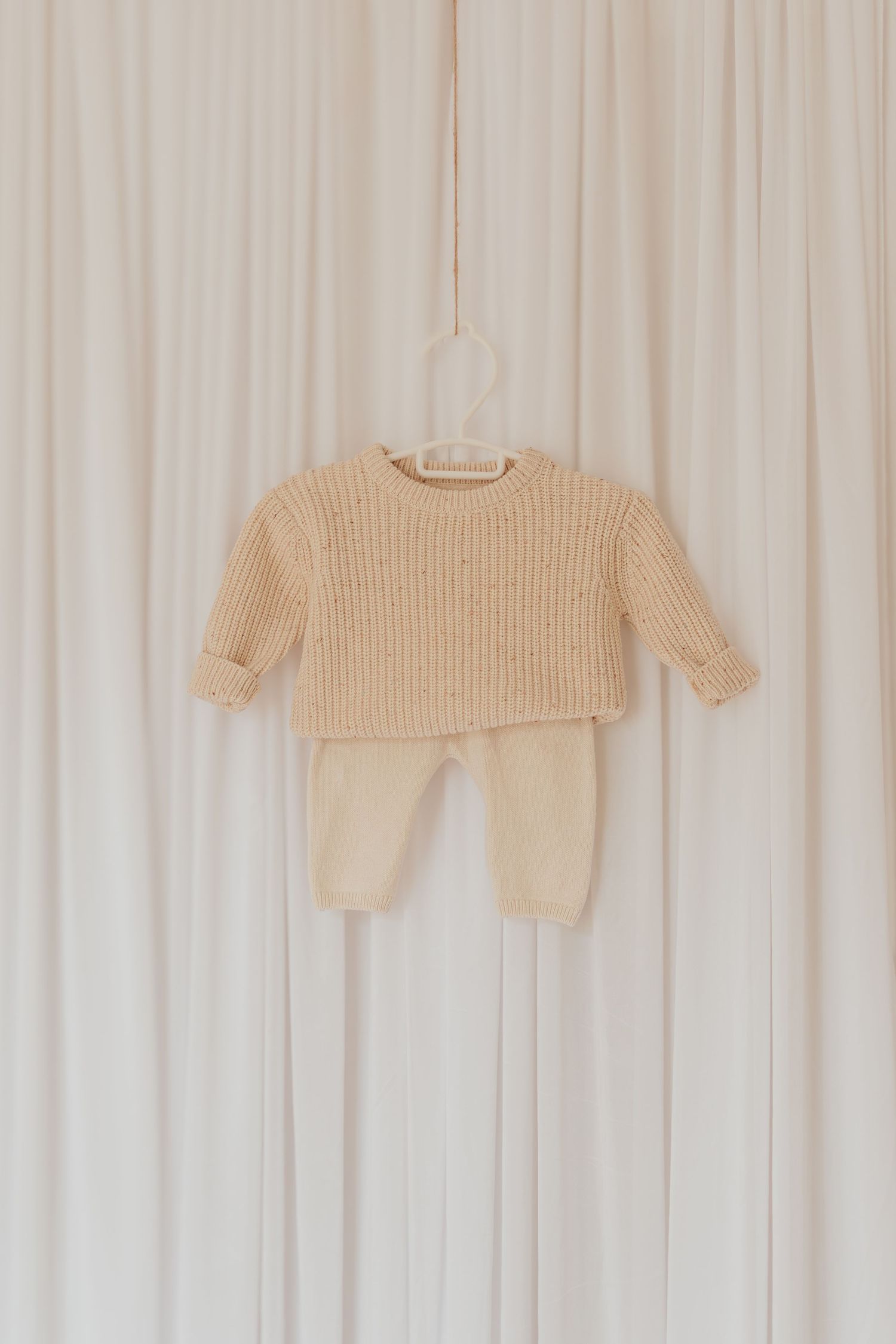 Cremefarbene Strick-Baby-Outfits, die an einer weißen Holzwand in einer minimalistischen Kinderzimmer-Umgebung hängen.