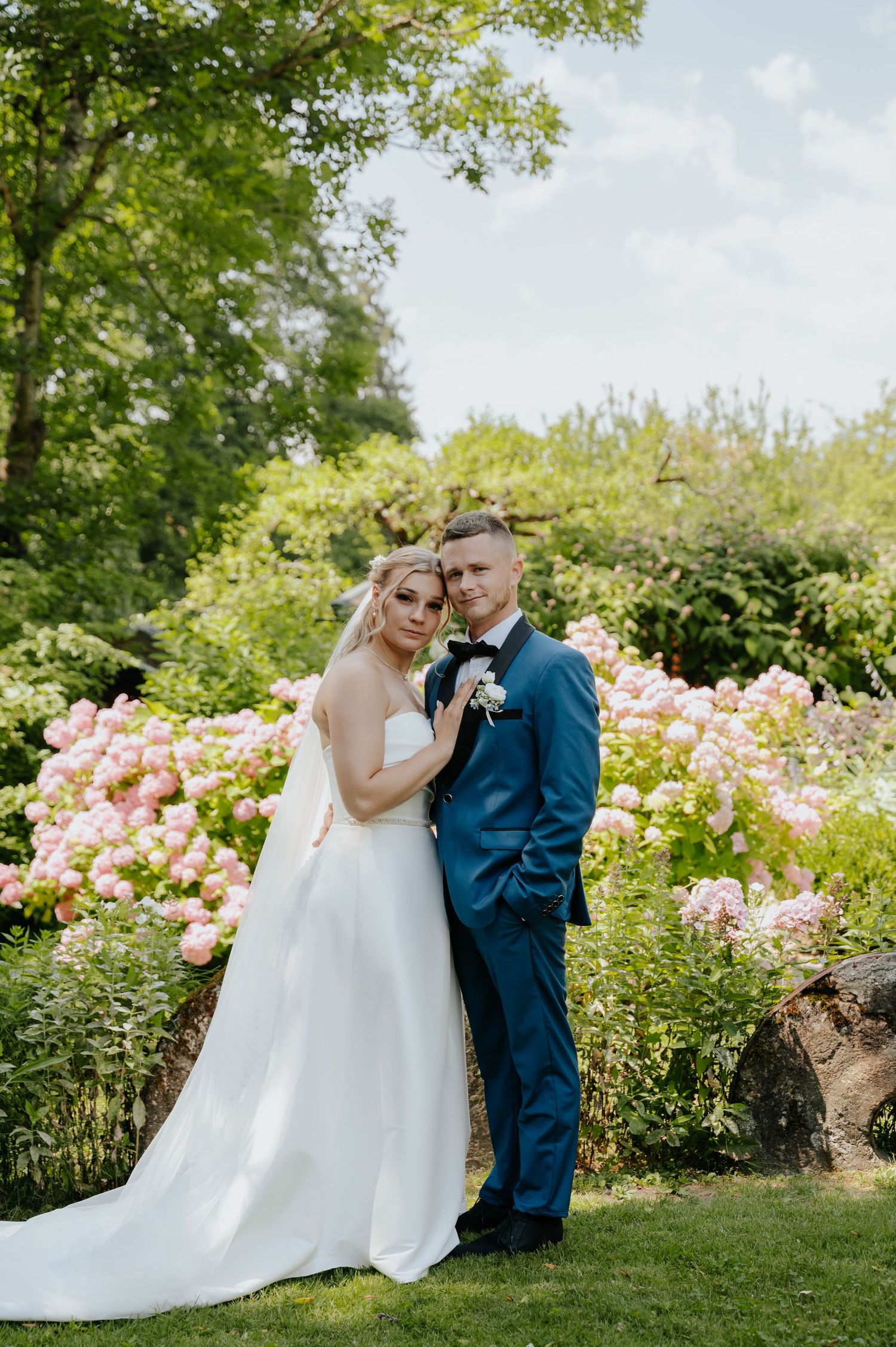 Bride in white gown and groom in blue suit pose together in a lush garden with pink hydrangeas blooming behind them.