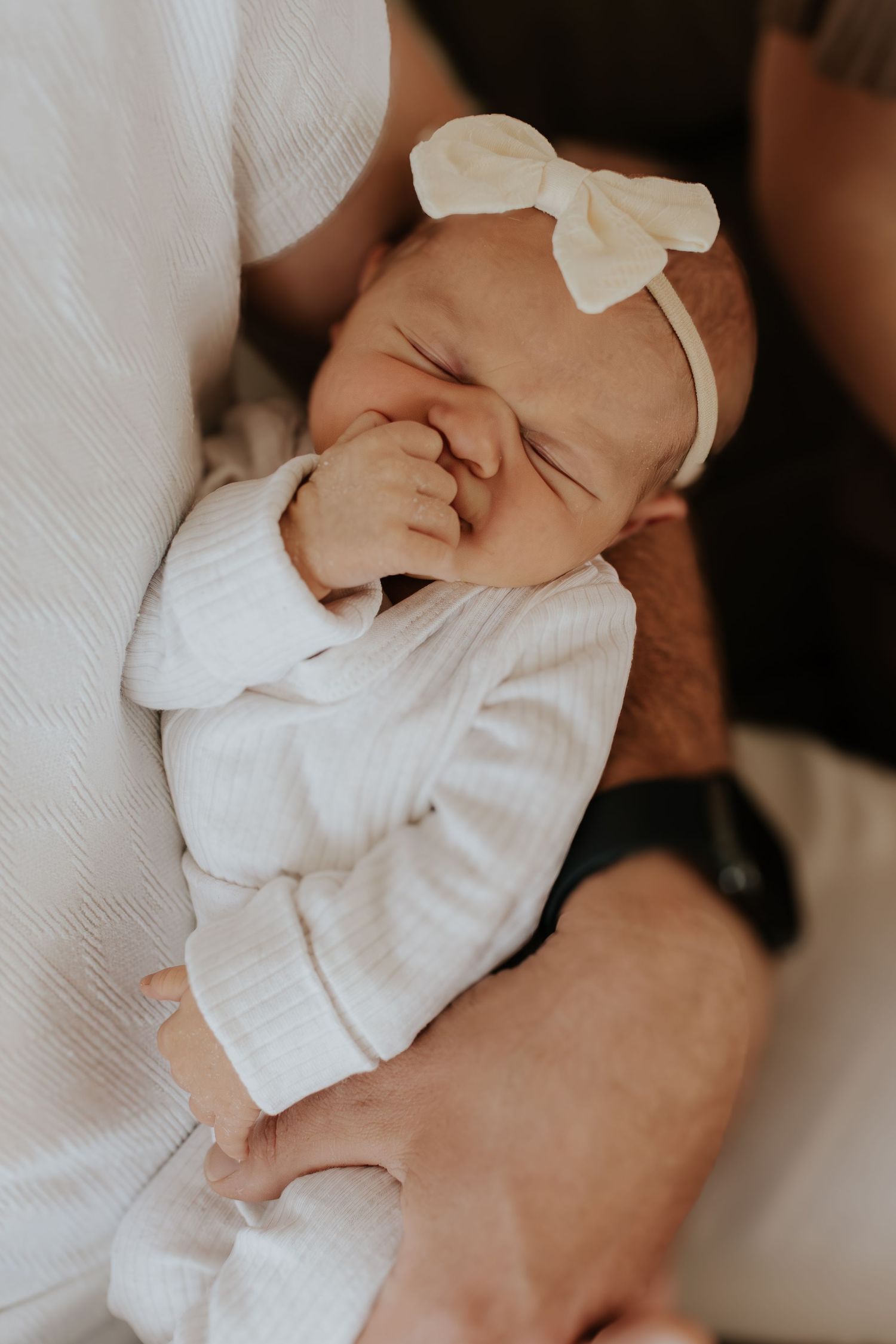 Newborn baby in white outfit and cream bow headband sleeping peacefully in parent's arms.