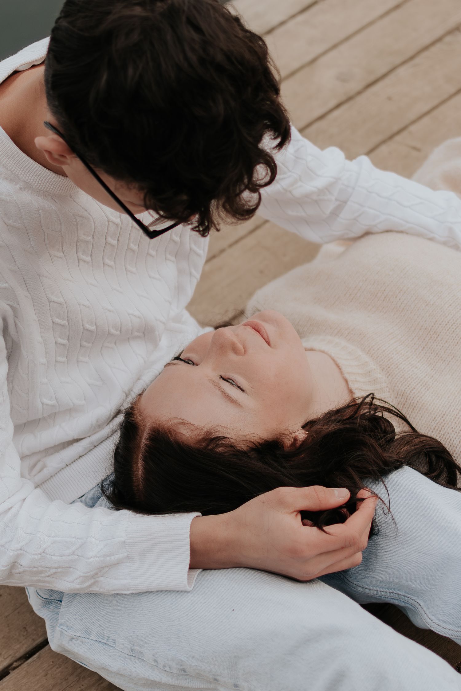 Two people in white sweaters share a tender moment on a wooden deck, one lying with eyes closed.