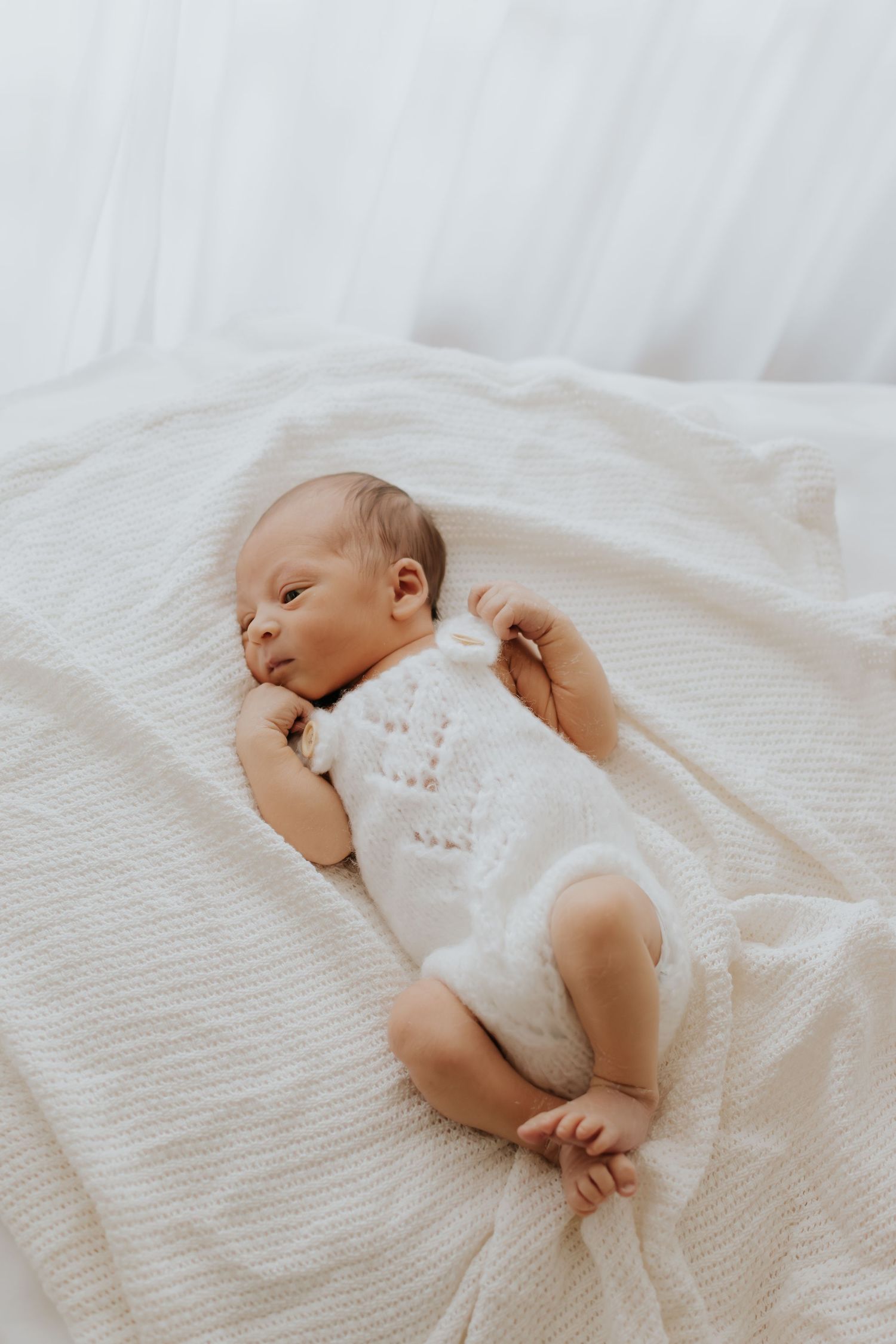 A sleeping newborn baby wearing white lace clothing rests peacefully on soft white bedding.