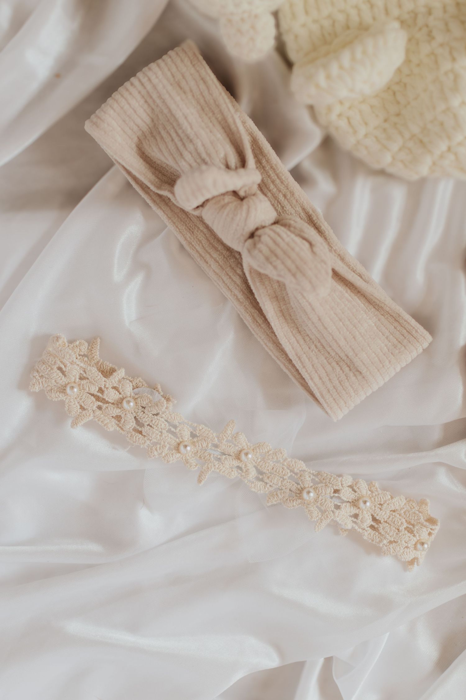 Beiges Strickstirnband mit Schleife und zartem Spitzenbesatz auf weißem Seidenstoffhintergrund.