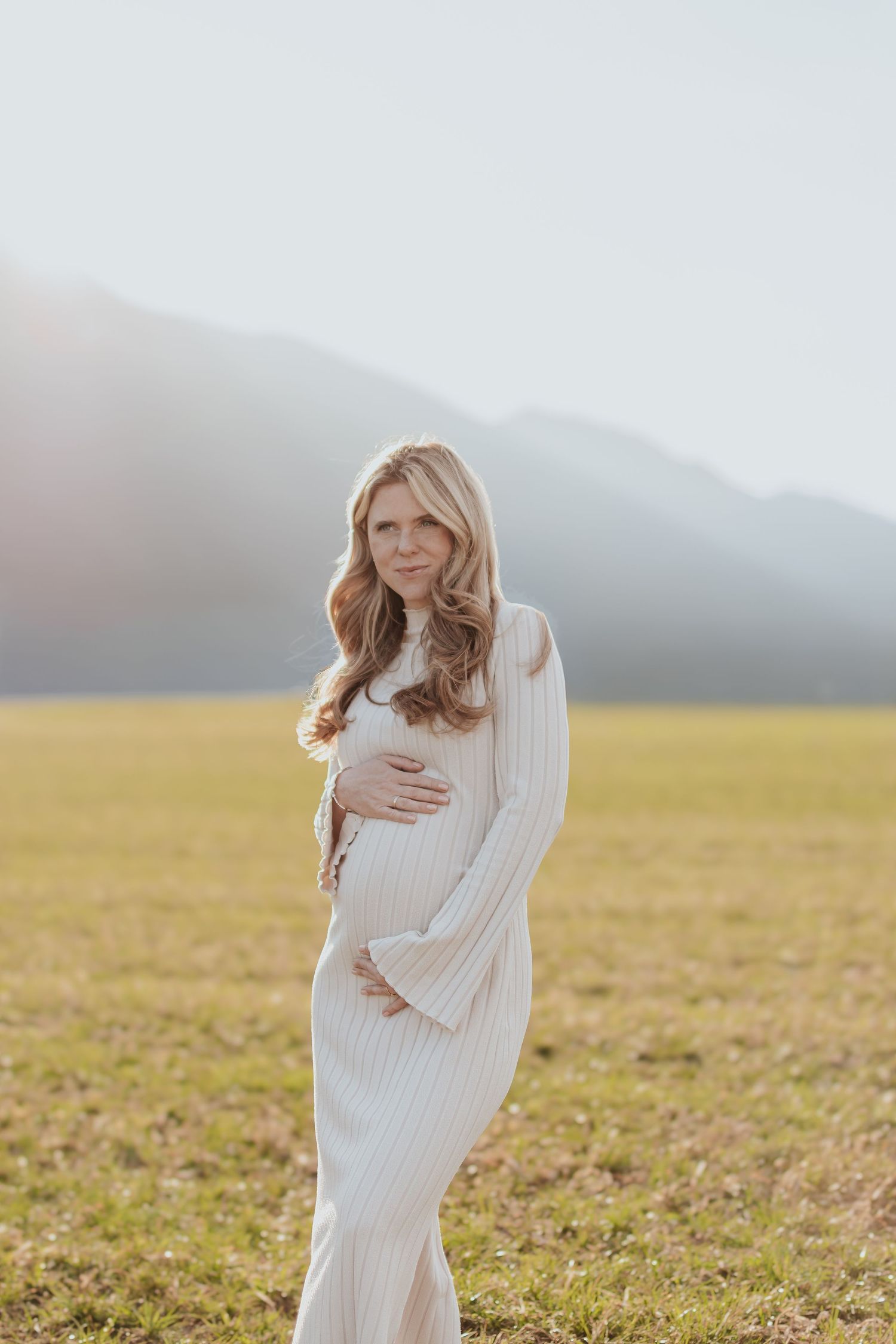 Pregnant woman in white ribbed maxi dress cradling baby bump in sunlit field with mountain backdrop.