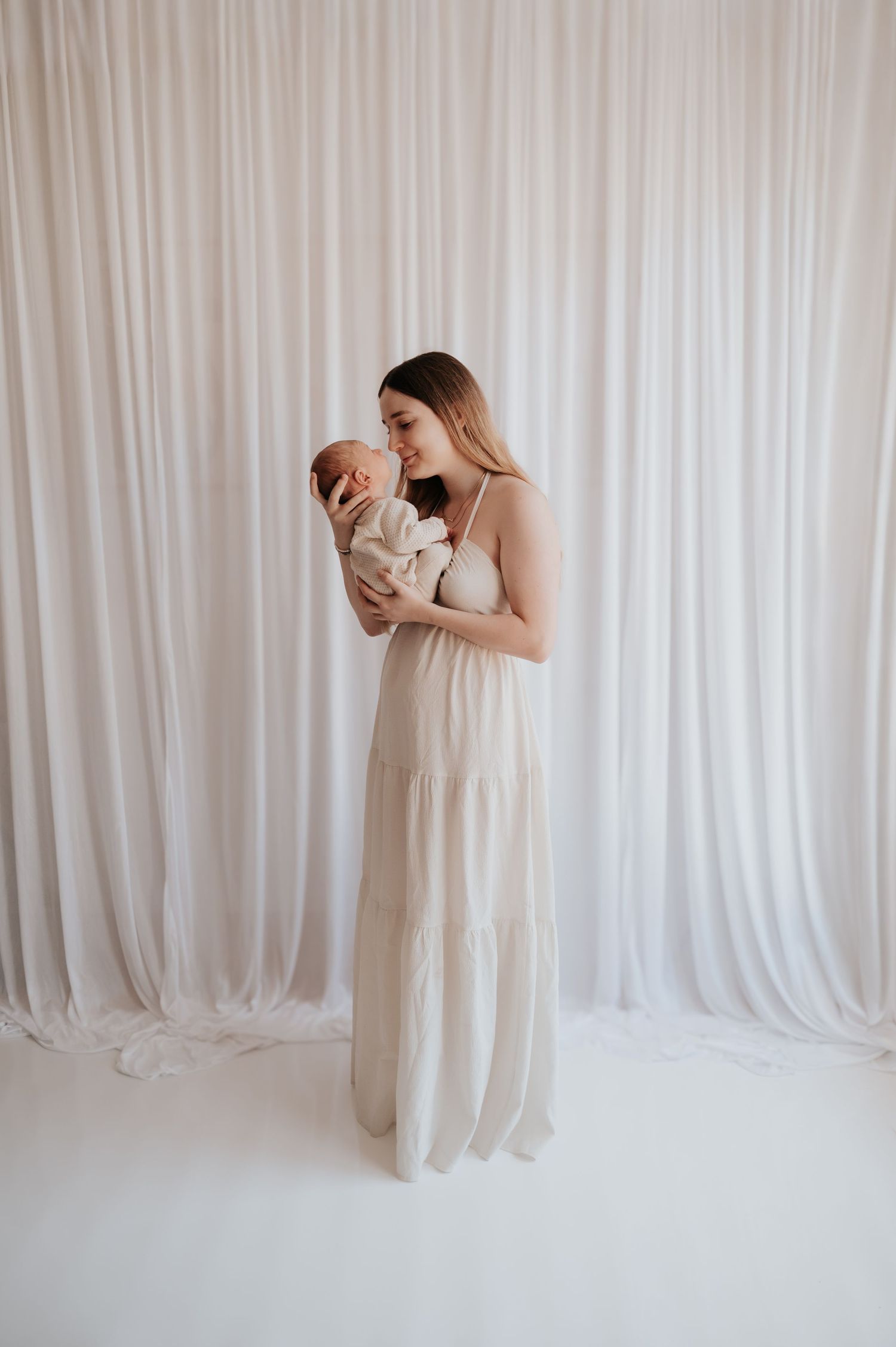 A mother in a white maxi dress tenderly holds her newborn baby against a soft white curtain backdrop.
