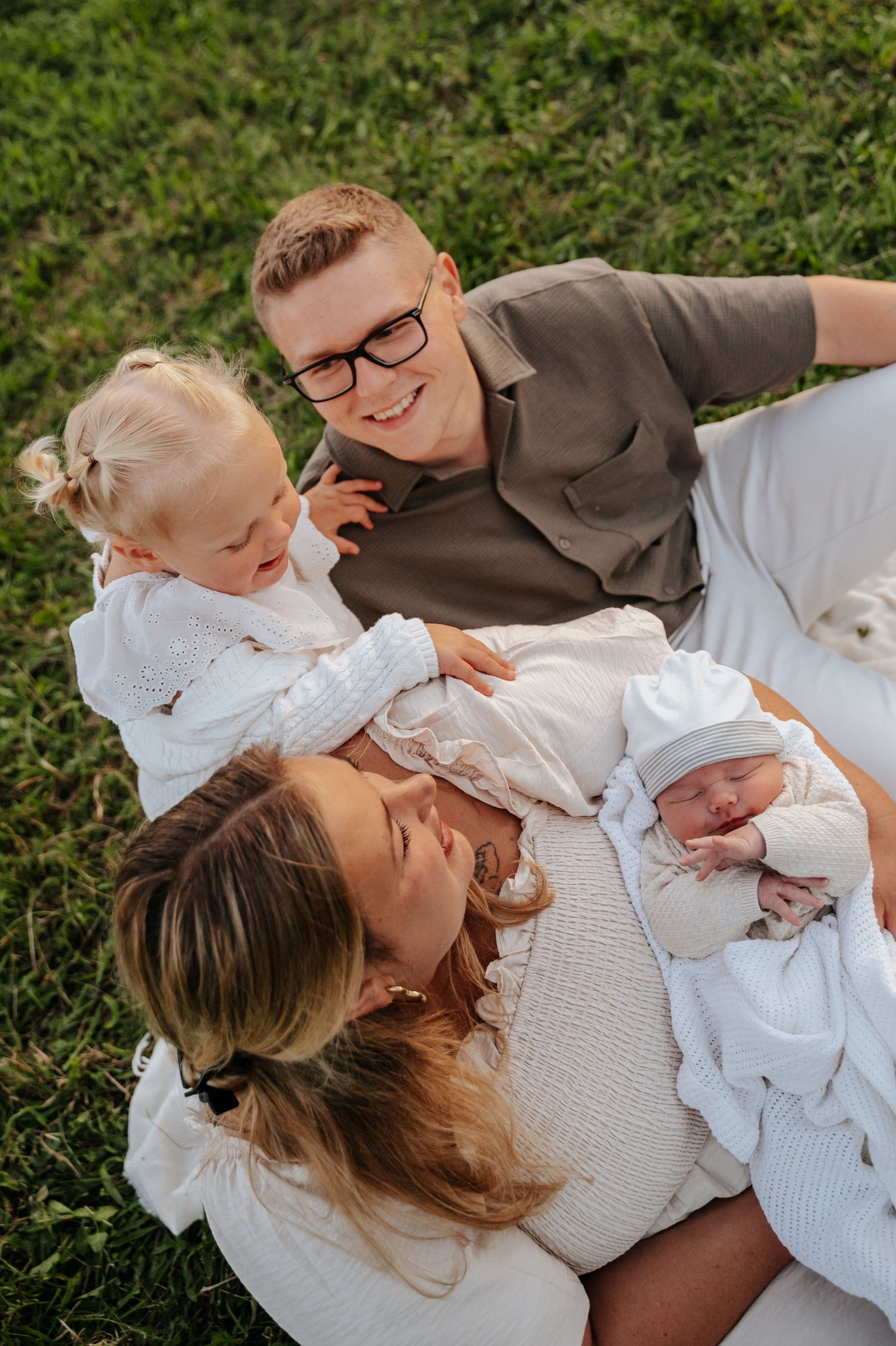 Family of four sitting on grass, parents smiling at newborn baby while toddler girl sits nearby.