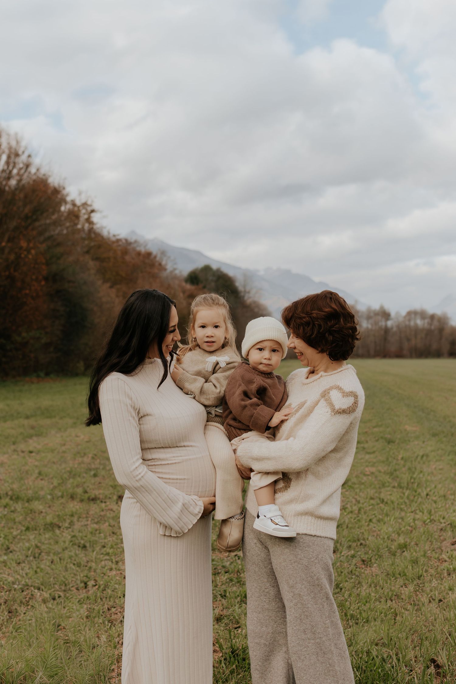 Family wearing neutral colored sweaters embrace lovingly in a grassy autumn field under cloudy skies.