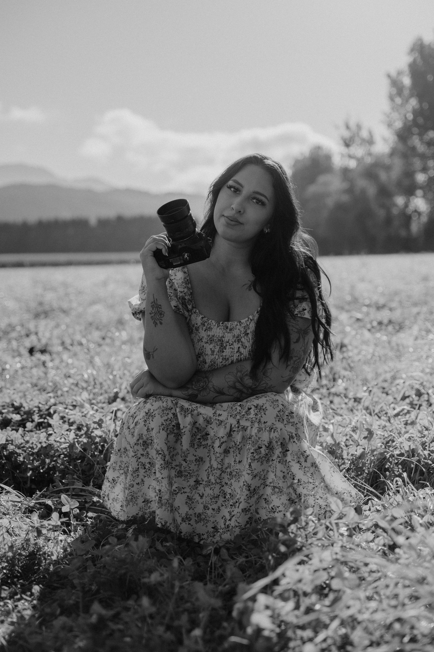 Black and white photo of a young woman in a floral dress holding a camera while sitting in an open field.