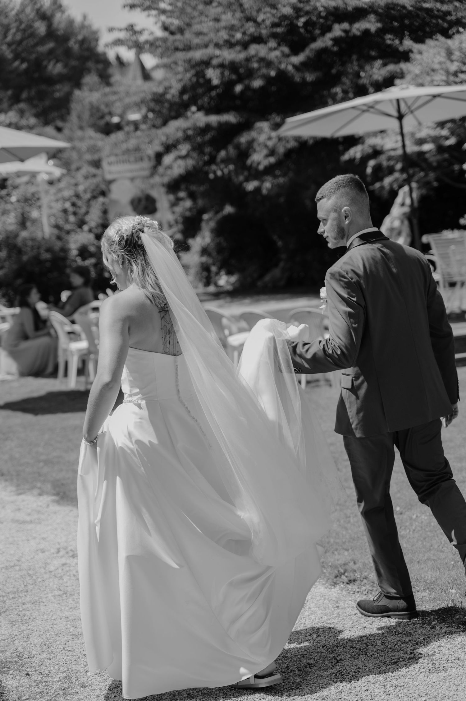 Black and white photo of a groom helping his bride with her flowing veil and gown as they walk outdoors.