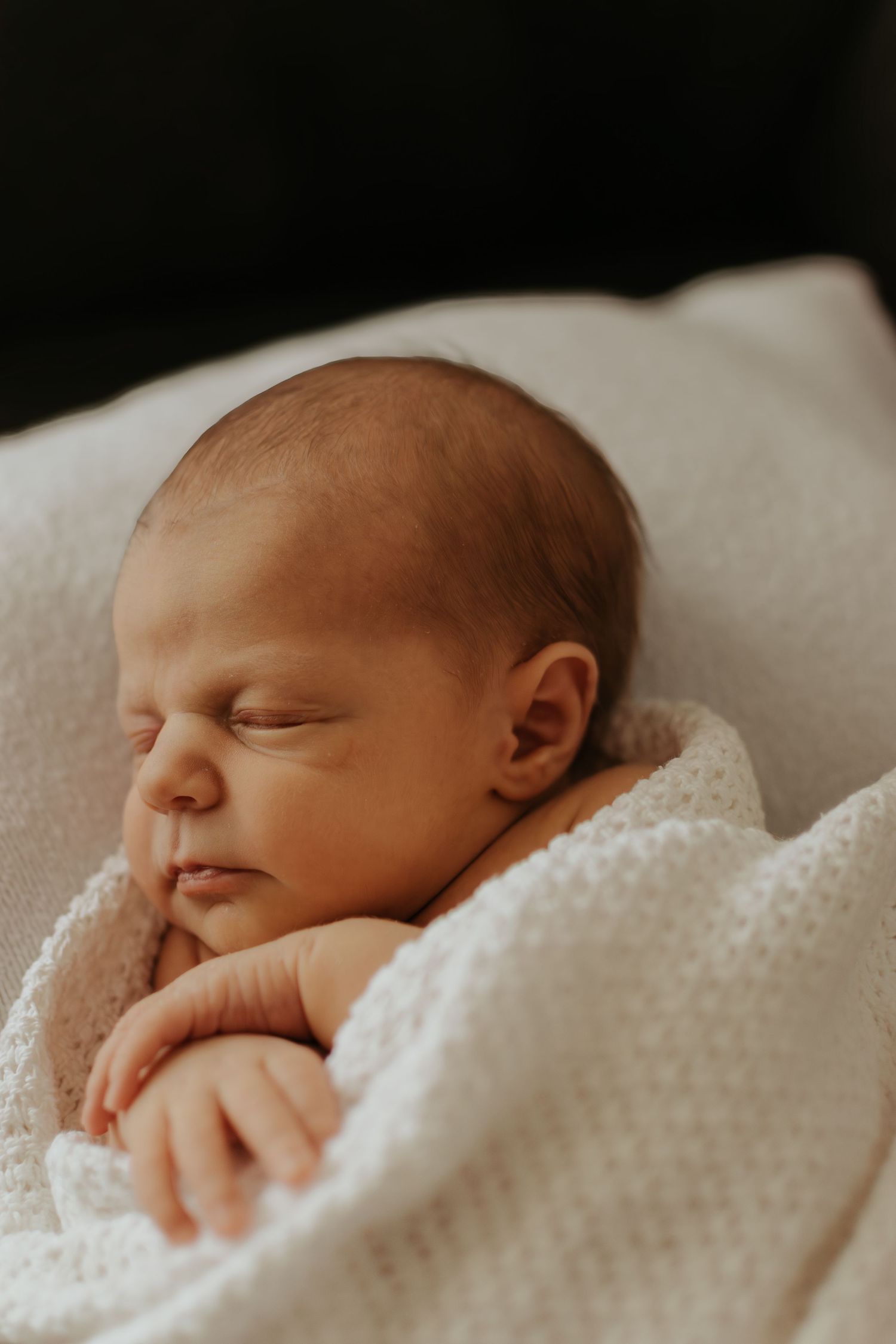 A peaceful newborn baby sleeping soundly wrapped in a soft white knit blanket with tiny hands visible.