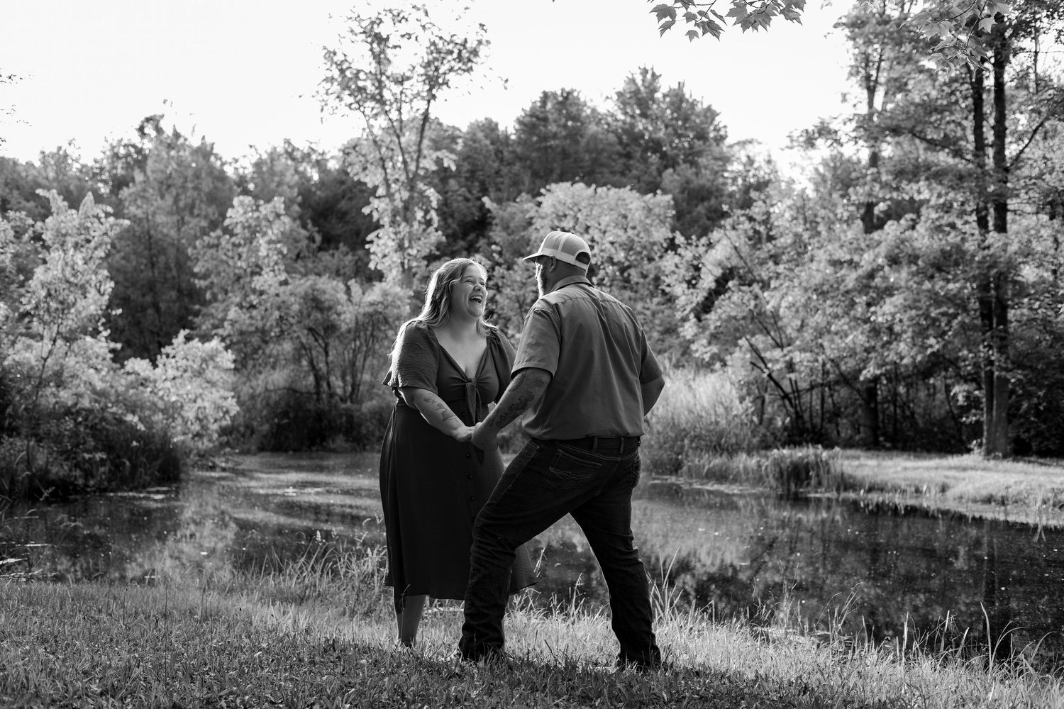 Black and white photo of a couple dancing together near a pond surrounded by lush trees outdoors.