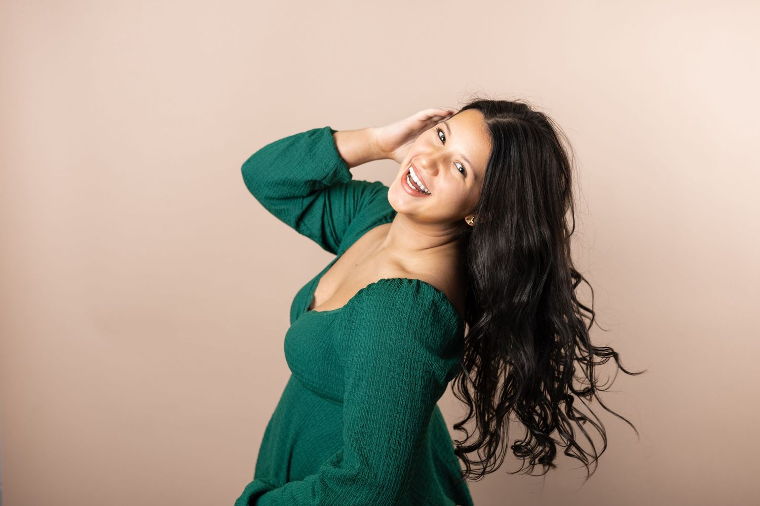 Person with long dark hair wearing an emerald green sweater poses playfully against a beige background.