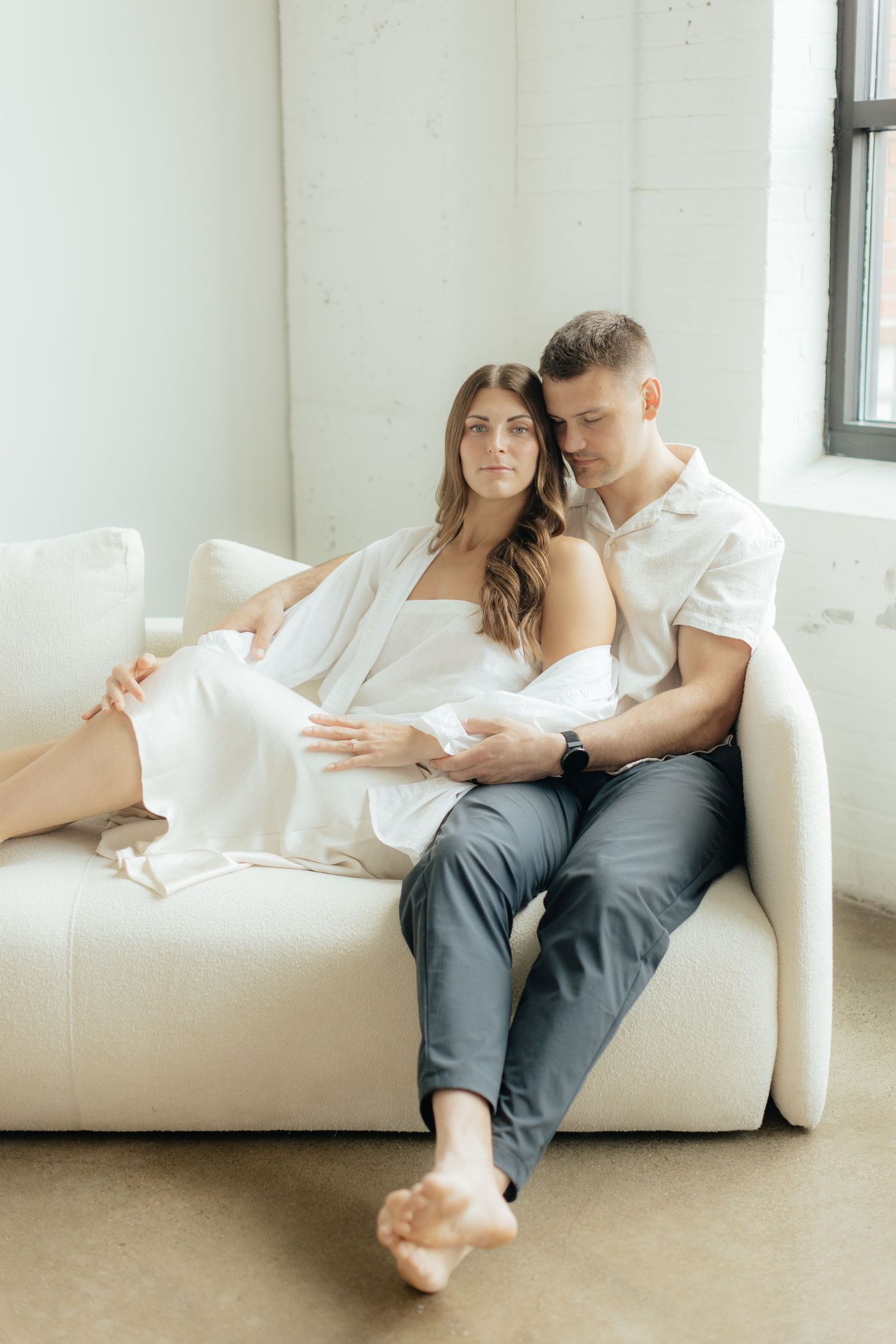 Couple embracing in soft natural light at Abide Studio in Minneapolis, MN, with minimalist white walls and warm, cozy tones