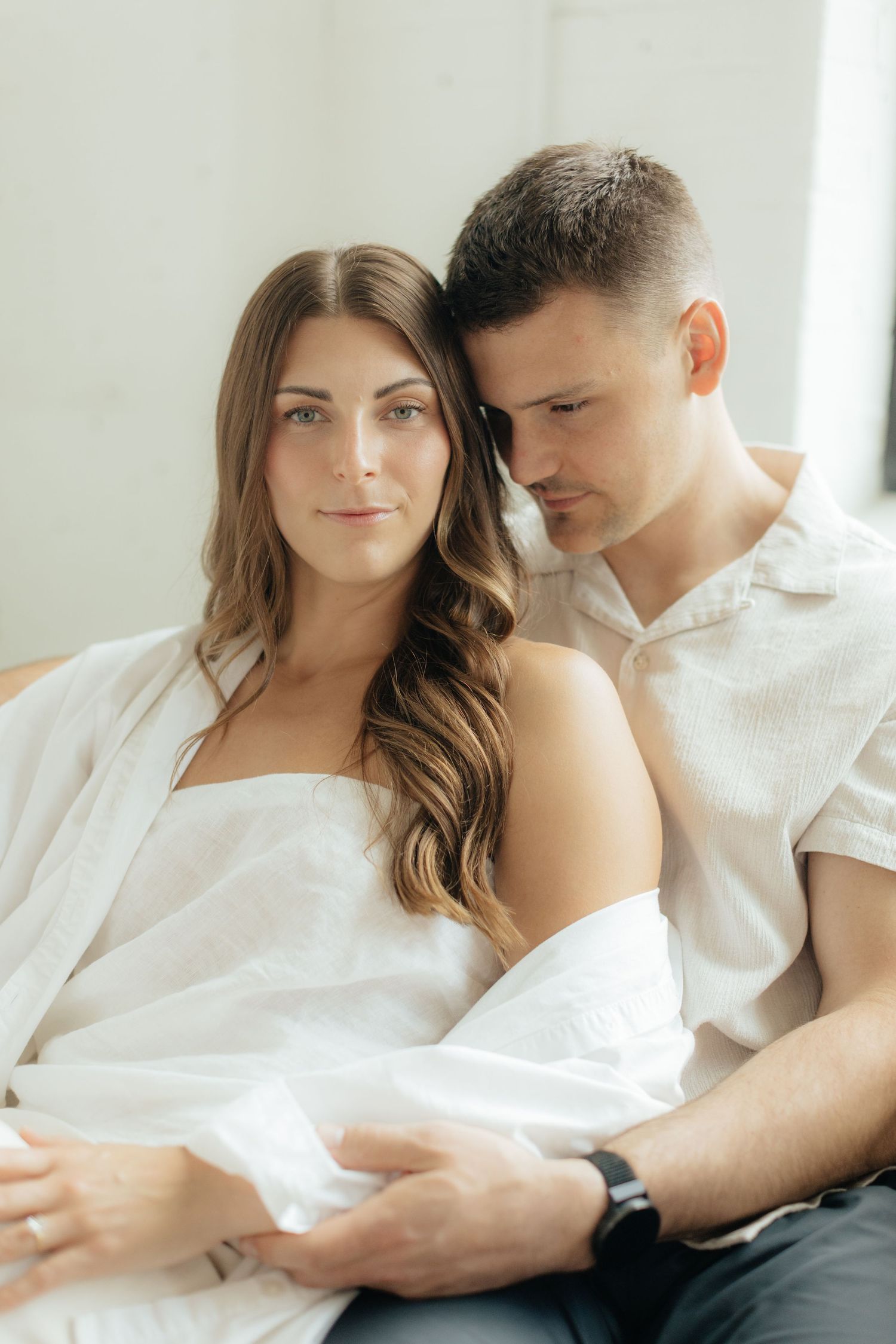 Couple embracing in soft natural light at Abide Studio in Minneapolis, MN, with minimalist white walls and warm, cozy tones