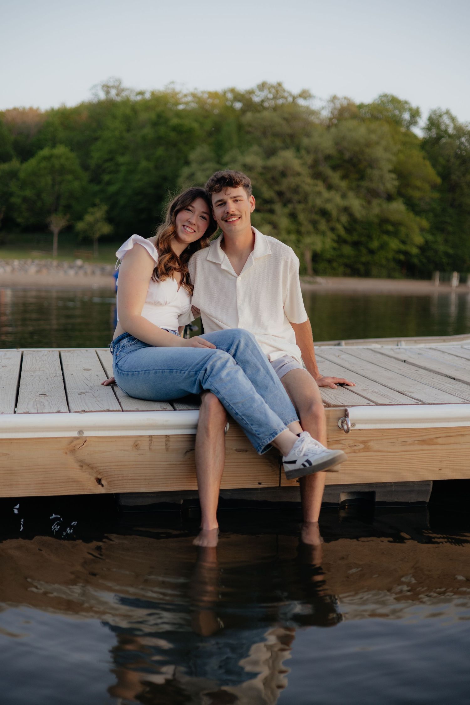Couple smiling together along the lakeshore at Fish Lake Regional Park in Maple Grove, MN, with bright green trees, clear blue water