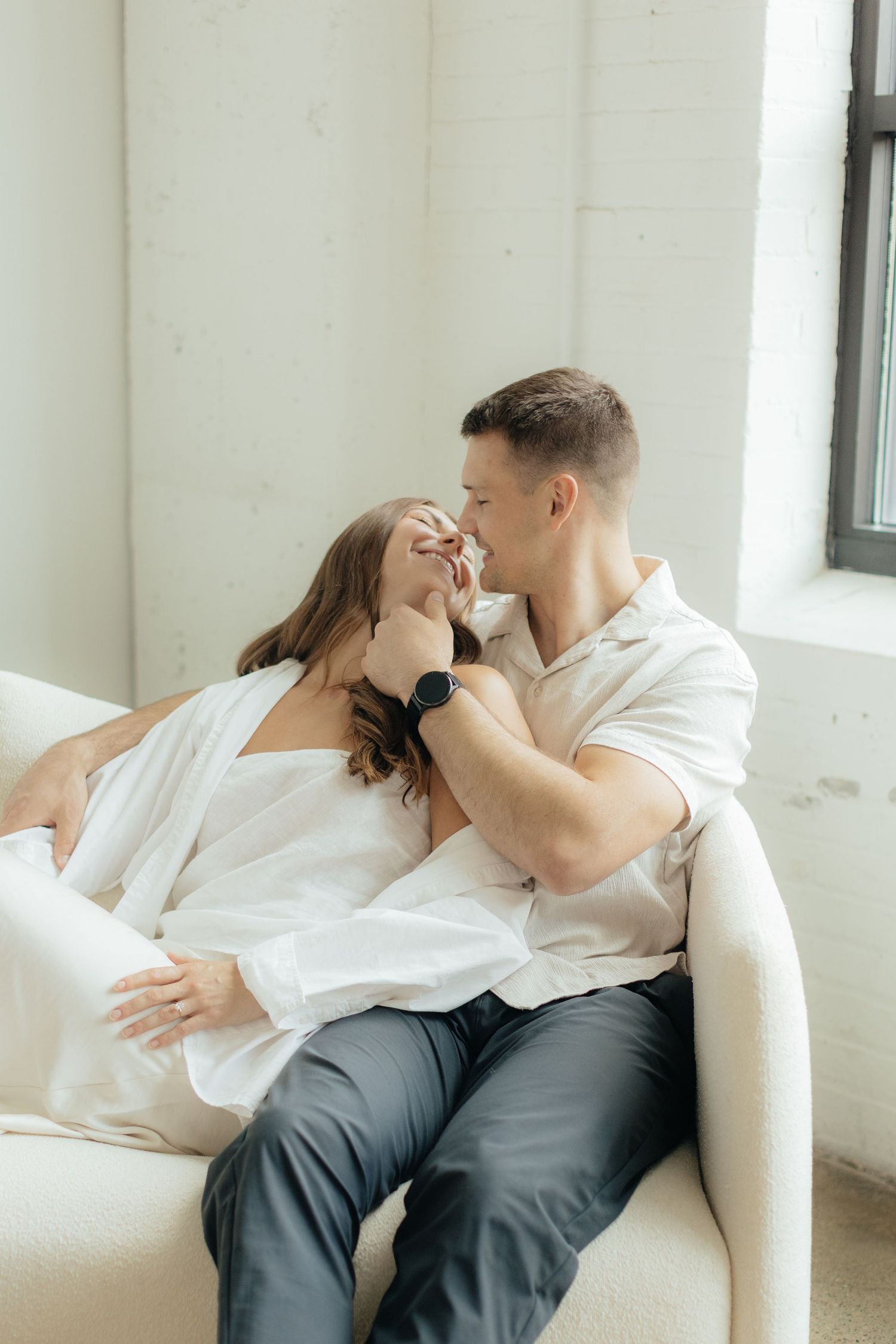 Couple embracing in soft natural light at Abide Studio in Minneapolis, MN, with minimalist white walls and warm, cozy tones