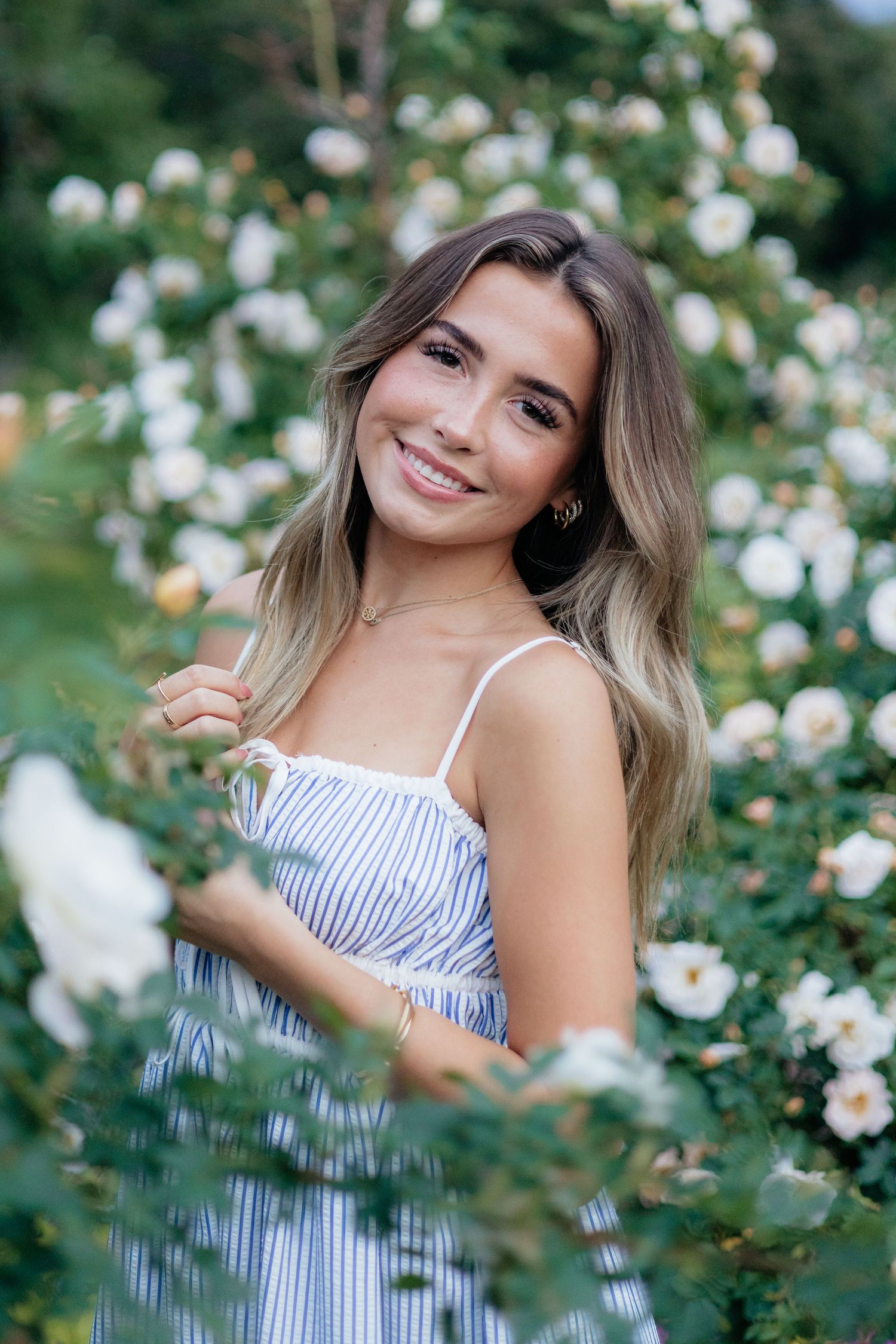 High school senior smiling and posing among blooming roses at Lyndale Park Rose Garden in Minneapolis, MN, surrounded by vibrant flowers and