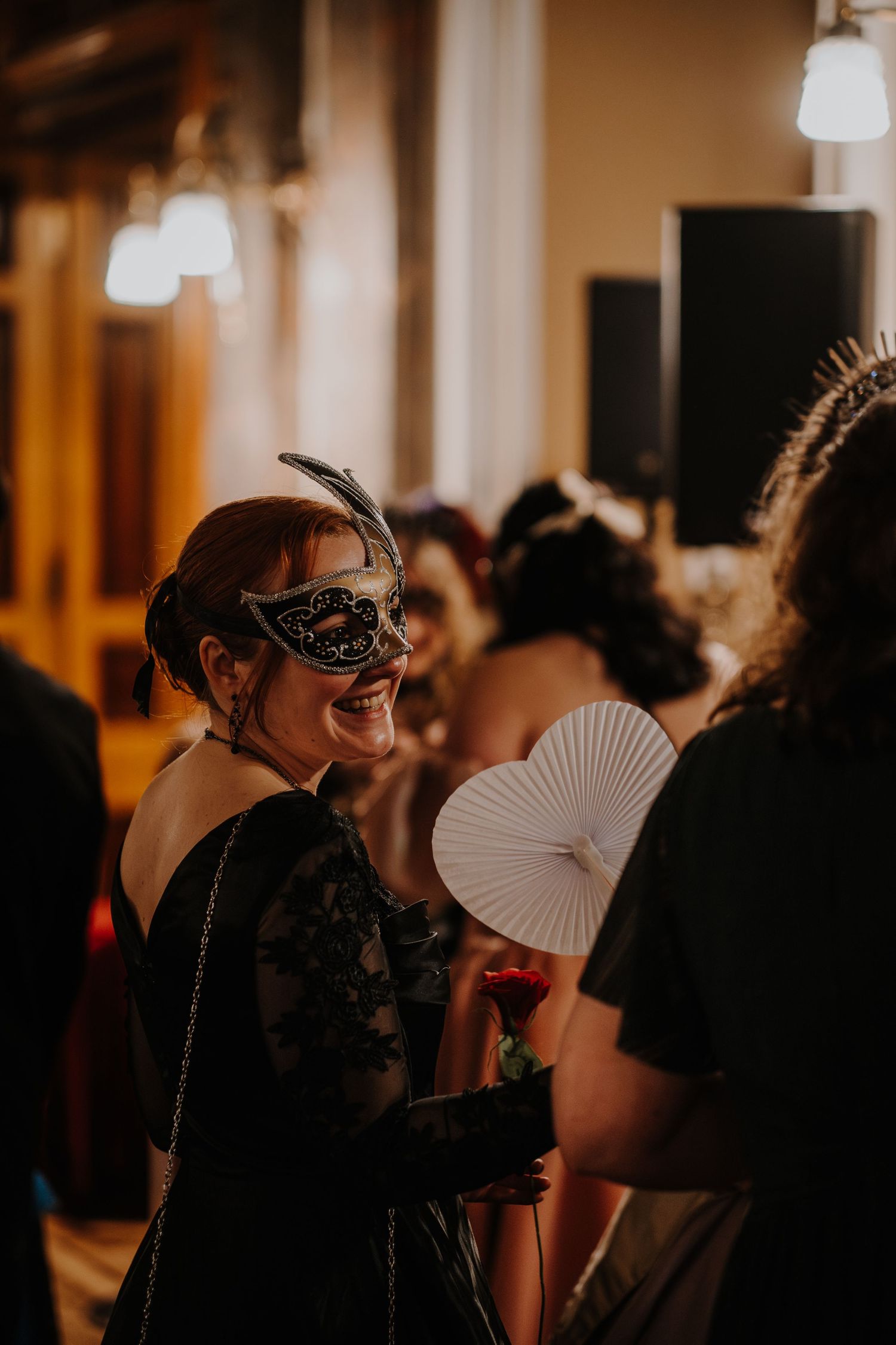 Guest in black dress and mask holds decorative fan at masquerade fantasy ball.