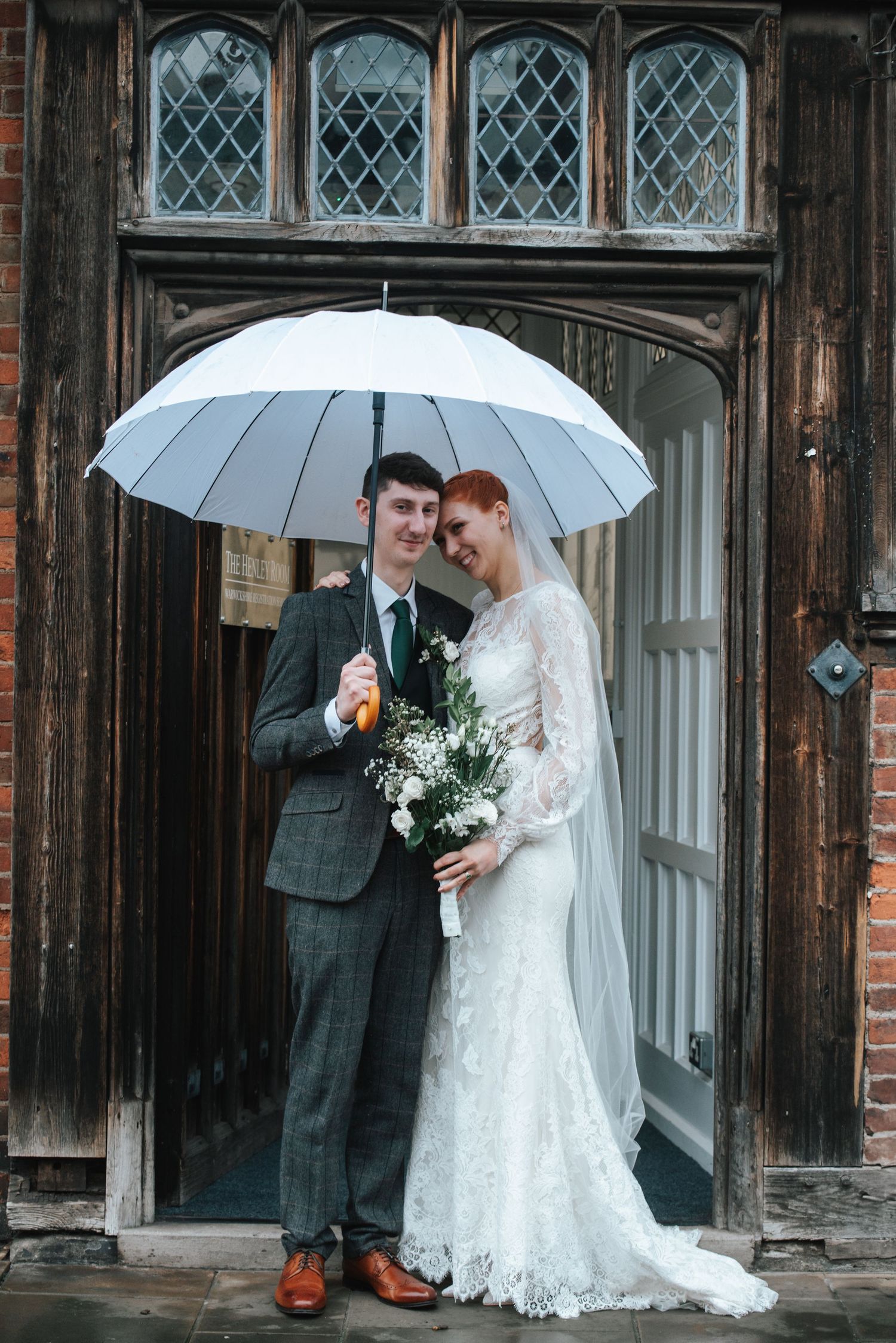 Newlywed couple shares intimate moment under white umbrellas against rustic wooden doors at historic venue.