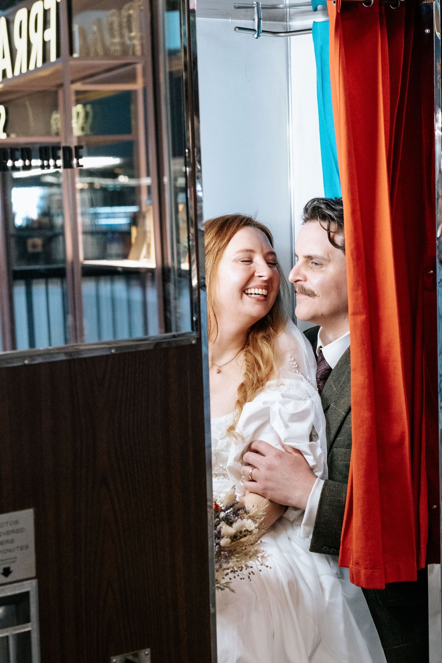 A laughing bride in a white dress holds flowers beside a suited groom in a vintage photo booth with red curtains.