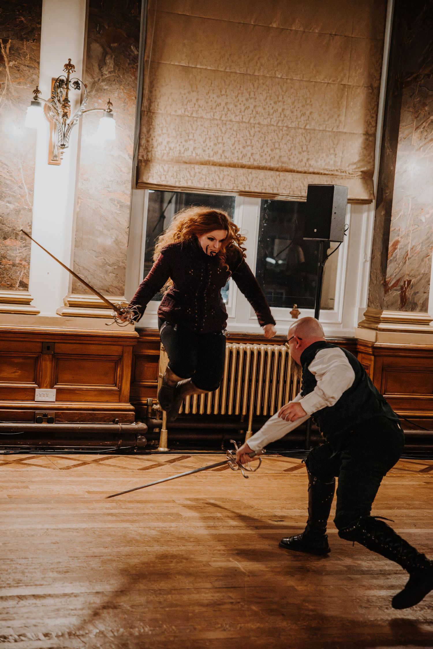 Two people practice sword fighting techniques in a dimly lit church or hall with wooden floors at a fantasy ball.