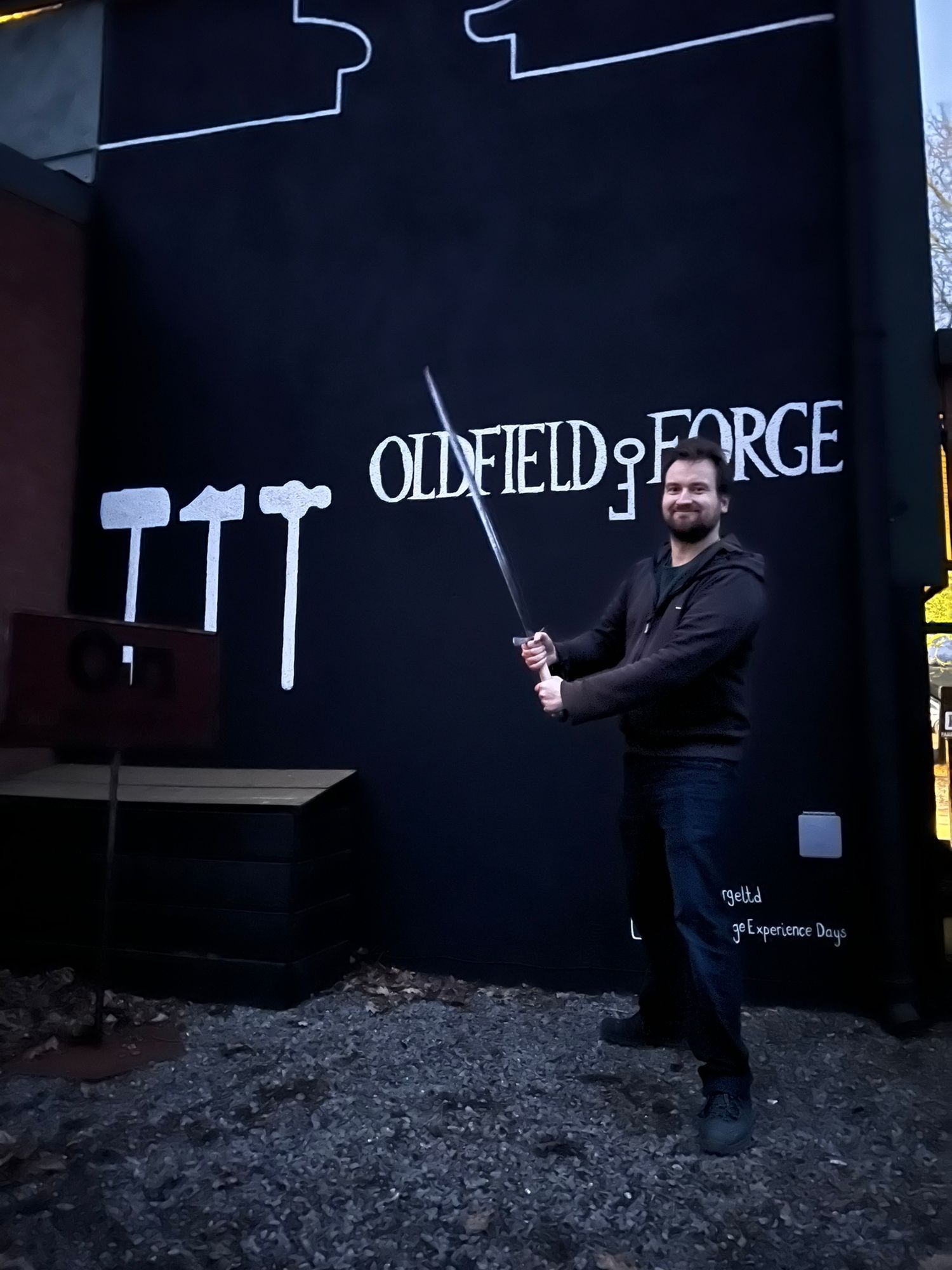 Person standing next to a black wall with 'Oldfield Forge' text and hammer designs.