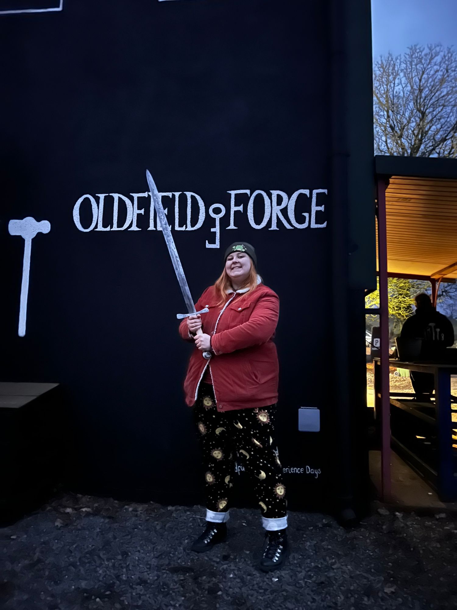 A person in a red coat stands against a dark wall with 'OLDFIELD FORGE' text visible in the background.