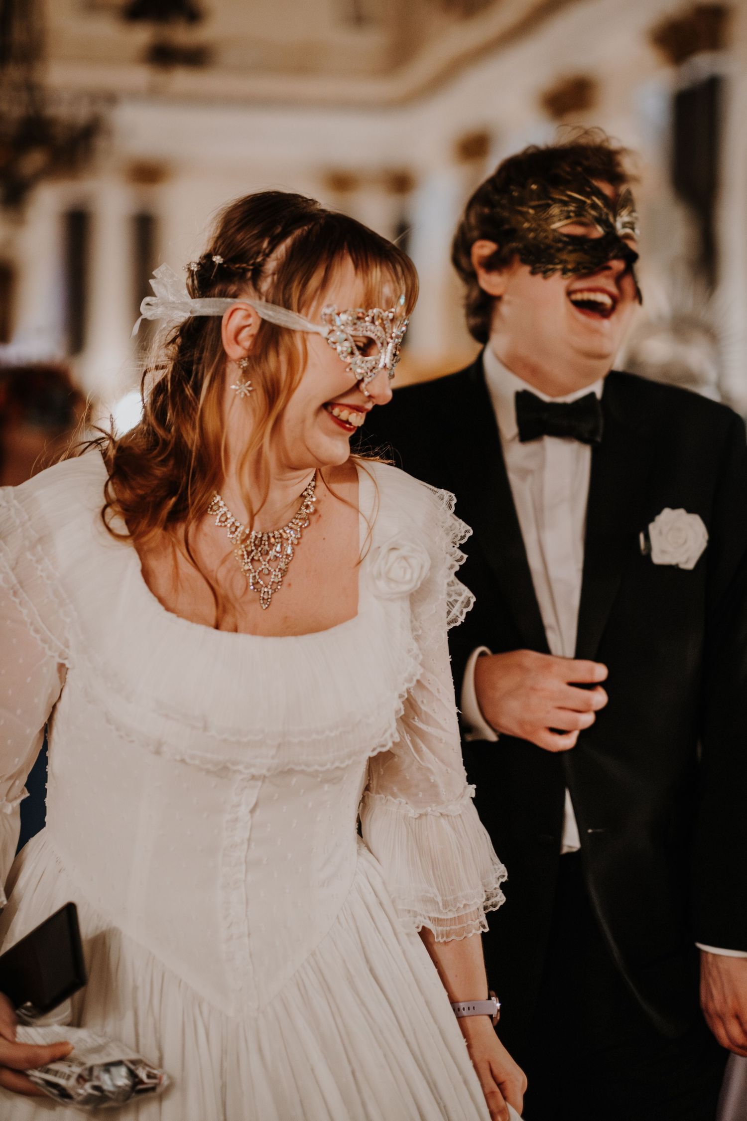 Couple in formal masquerade attire sharing a joyful moment at an elegant fantasy ball.