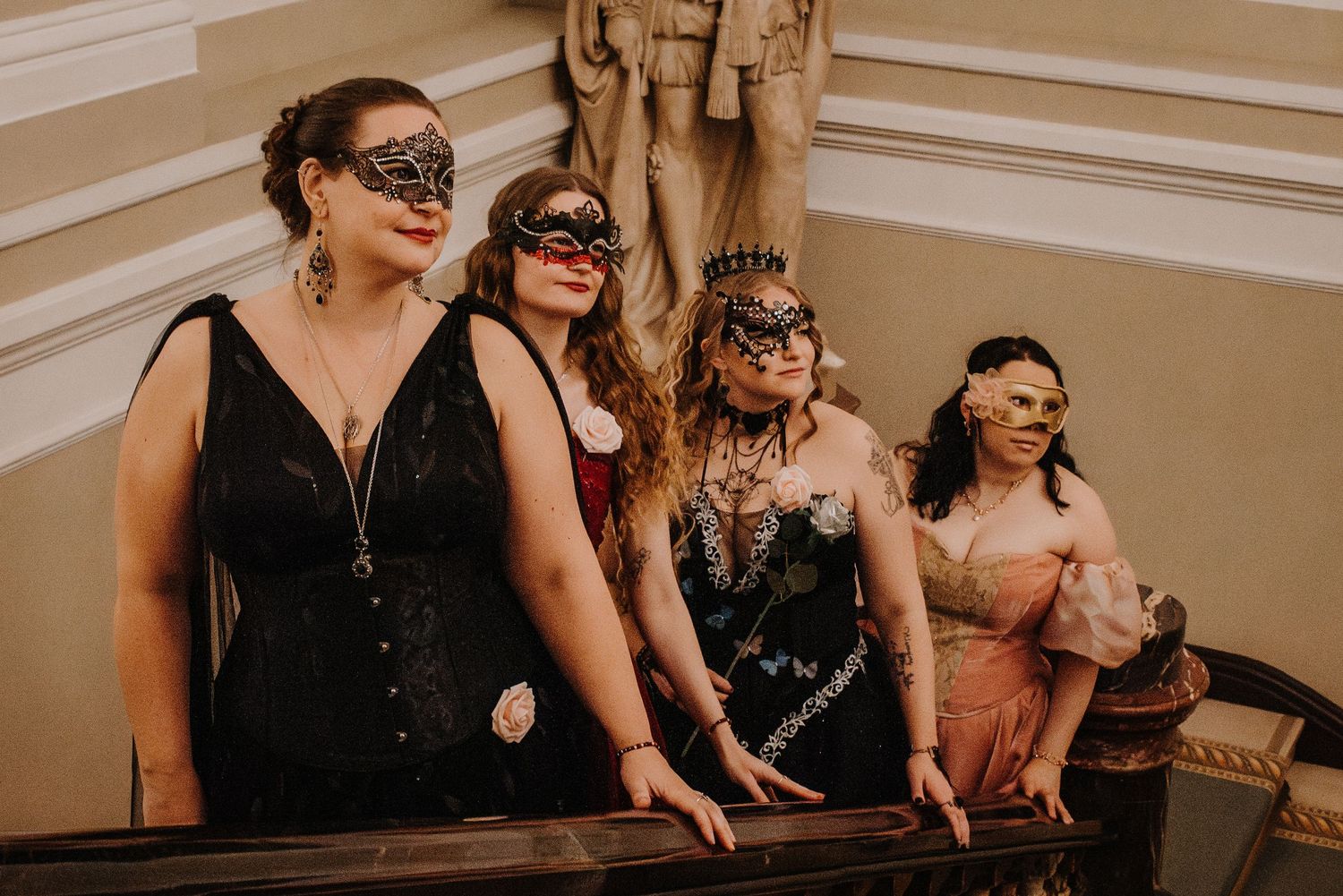 Group of guests in black formal wear and masks posing on elegant stairs at a fantasy ball.