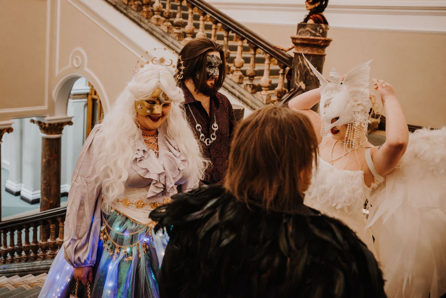 Costumed party guests in elaborate masks and formal wear gathering on an elegant staircase at a fantasy ball.