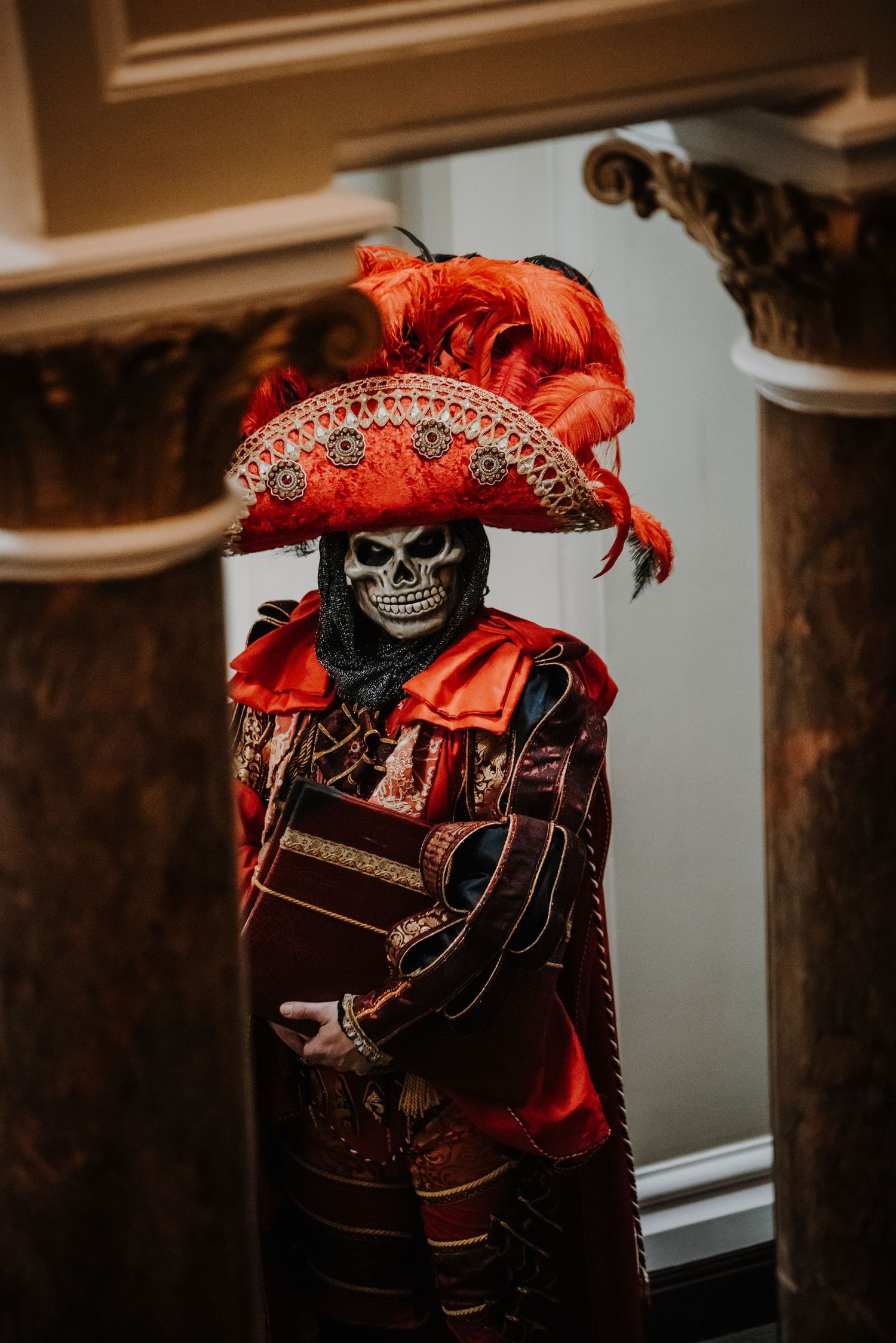 Skeleton figure in ornate red baroque costume with feathered hat at masquerade ball.