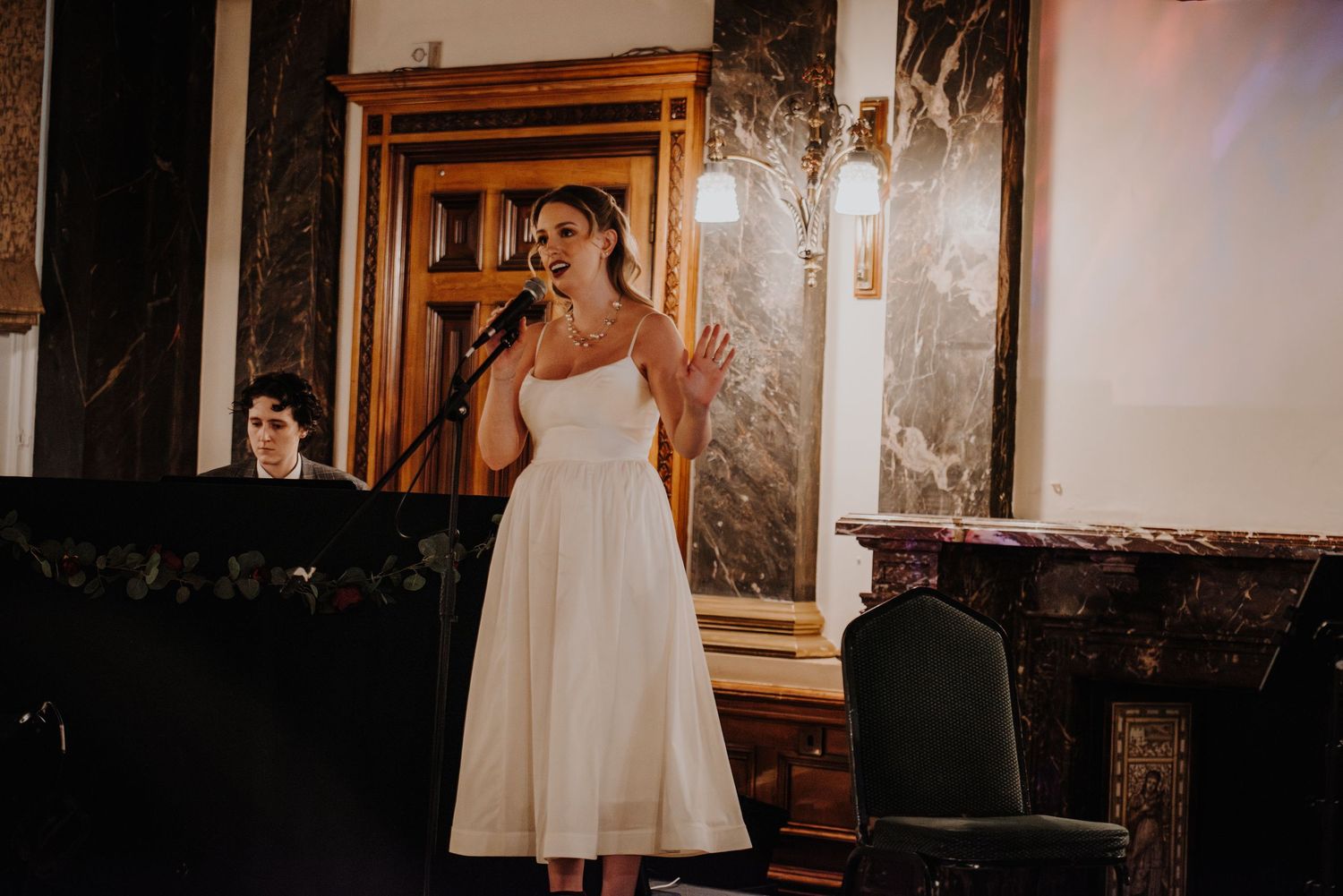 Performer in white dress stands at microphone against elegant wood-paneled wall at a fantasy ball.