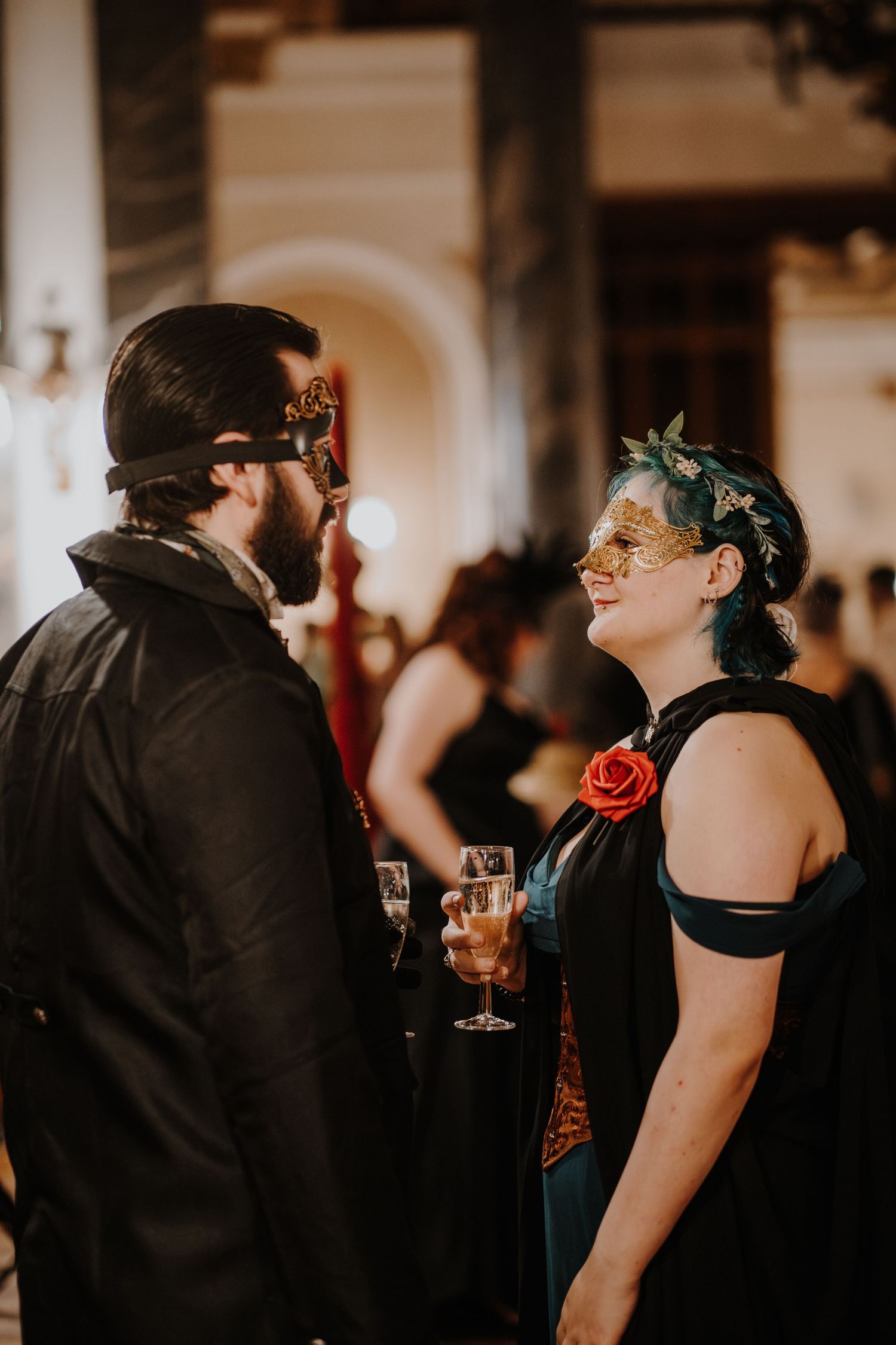 Guests in dark formal wear sharing a laugh while wearing decorative masks at a fantasy ball.