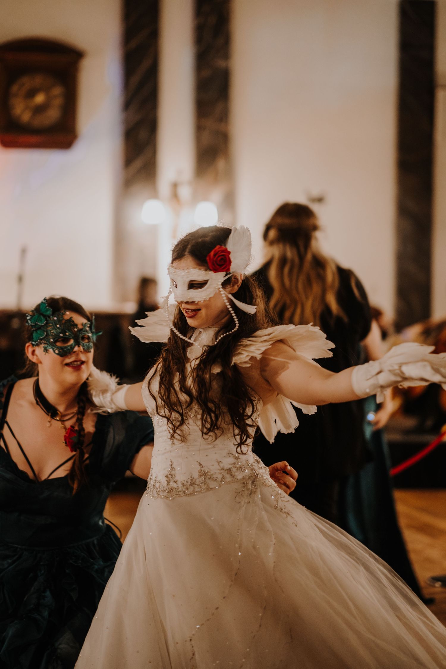 People wearing ornate masks dance together in  at a fantasy ball with warm lighting.