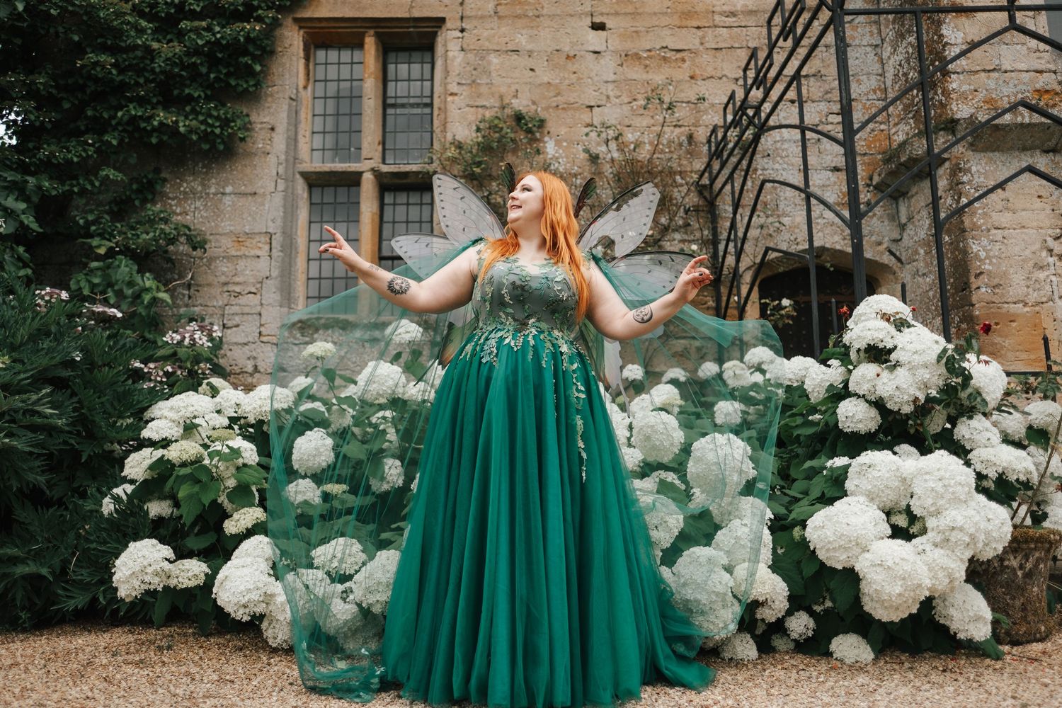 Person in emerald green ballgown posing among white roses against stone building.