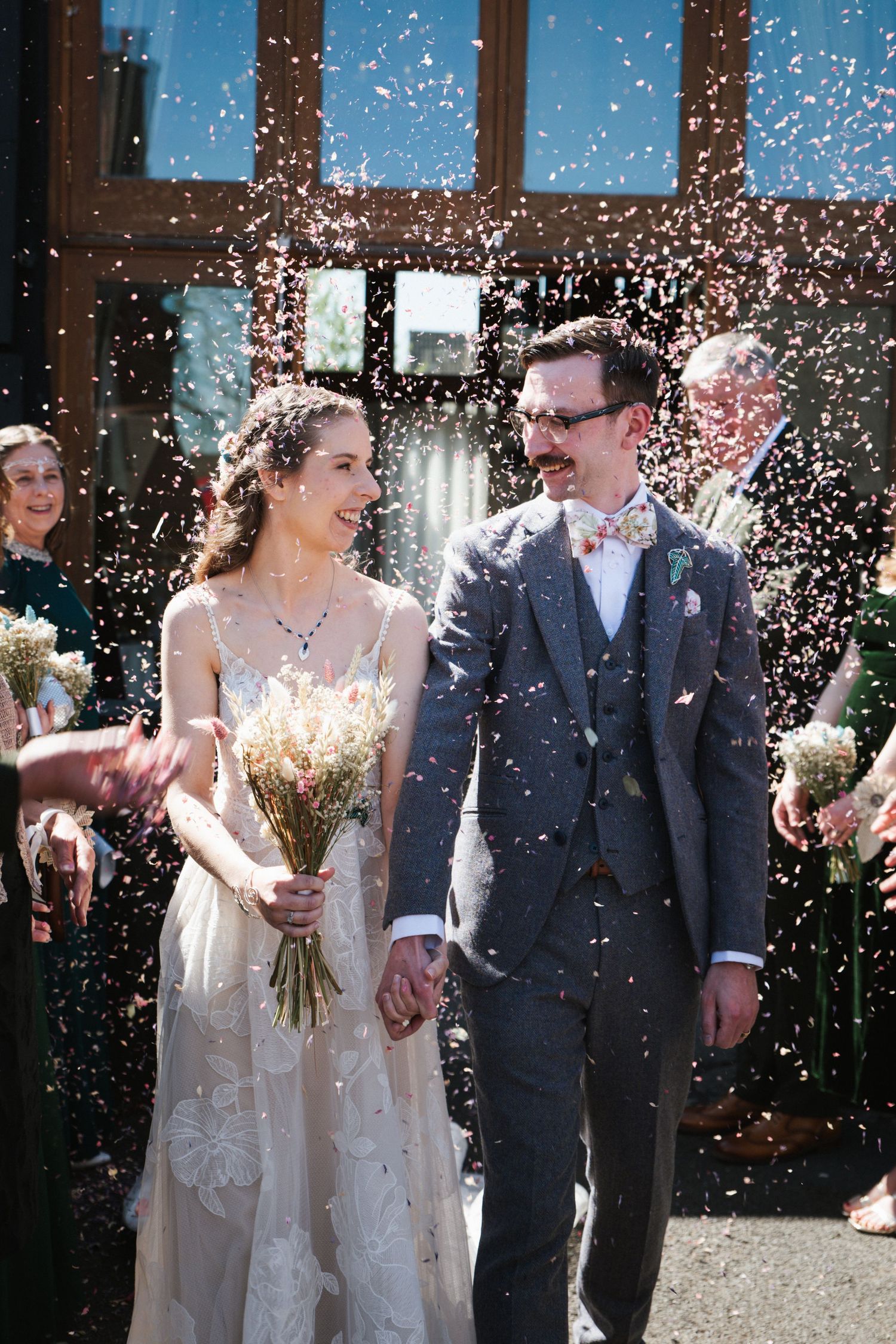 Newlywed couple walking through confetti shower, bride in white gown holding bouquet, groom in gray suit with bow tie.