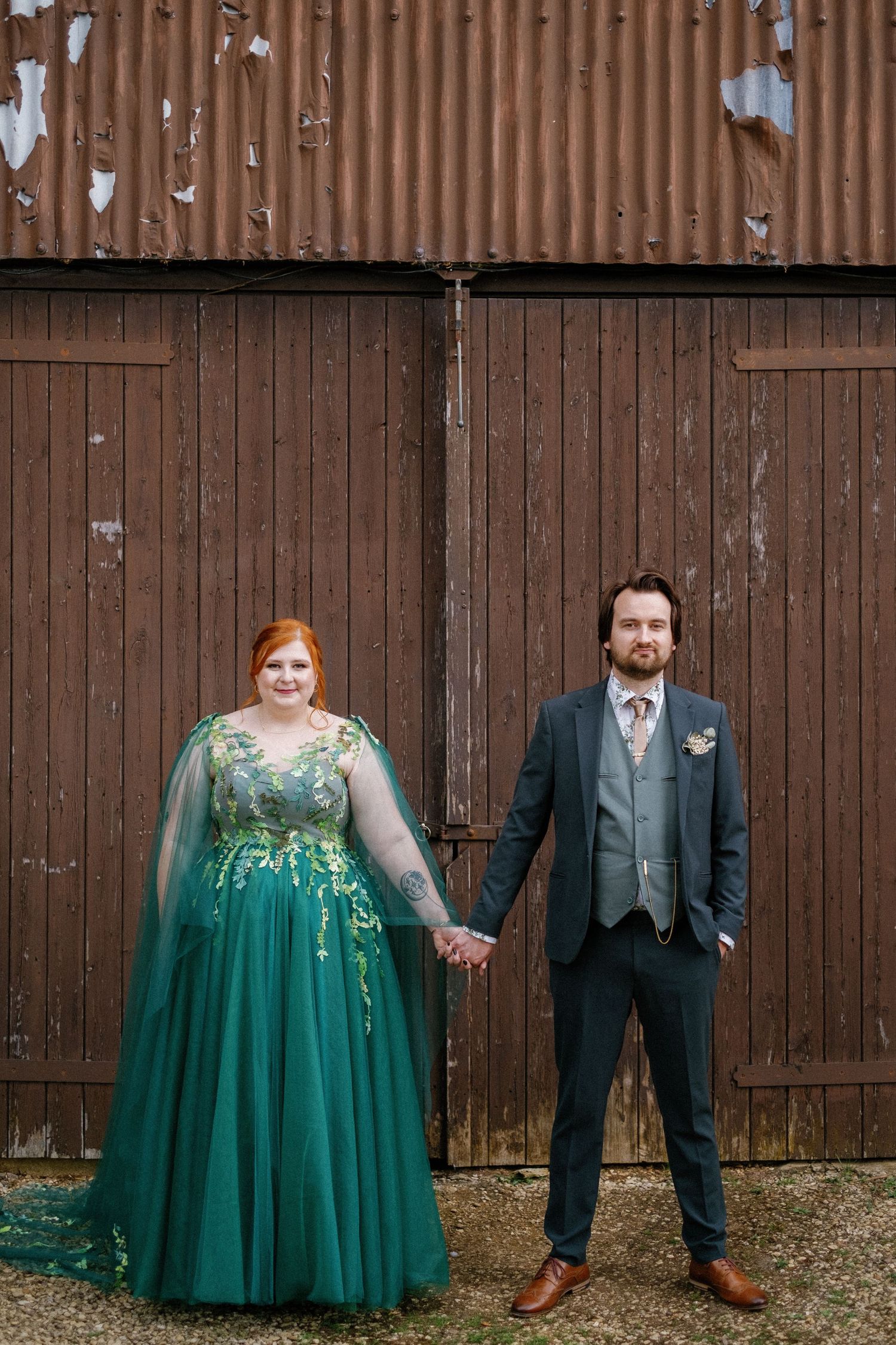 Couple posing against a rustic wooden barn wall, featuring an emerald green gown and gray suit.