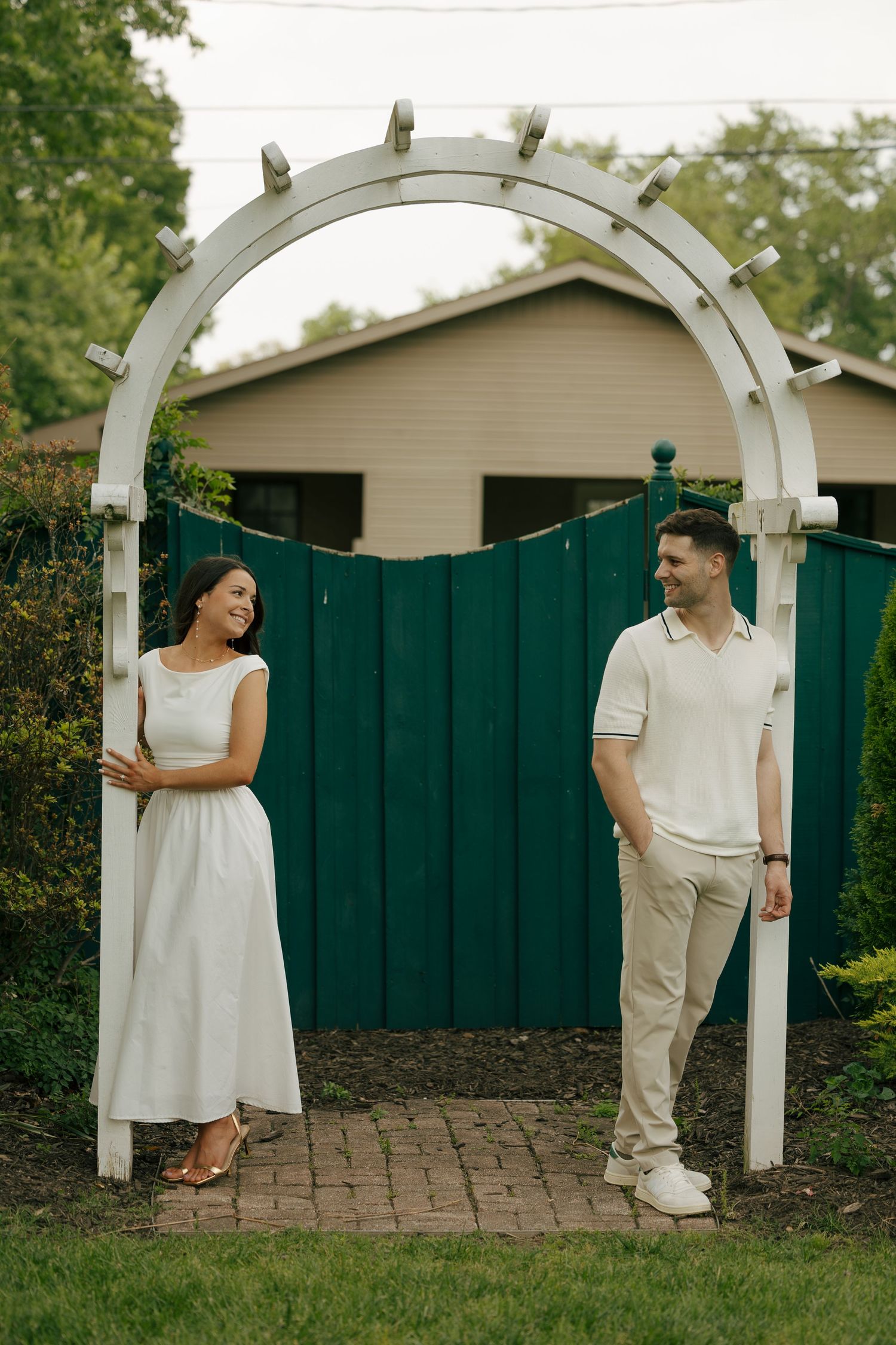 A couple smiles at each other under a white garden arch with a green fence backdrop.
