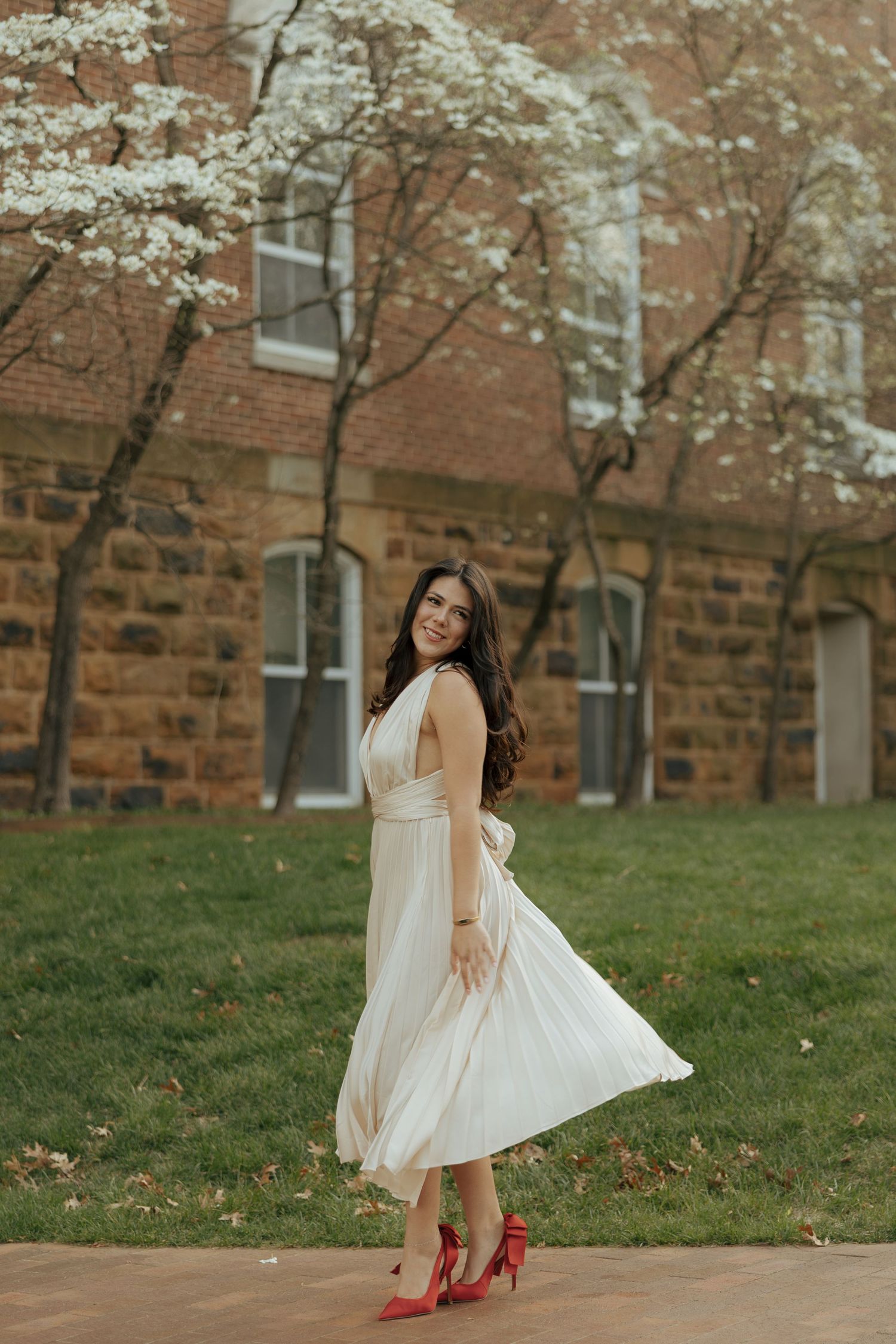 Woman in white midi dress and red heels twirling near flowering trees outside a brick building.