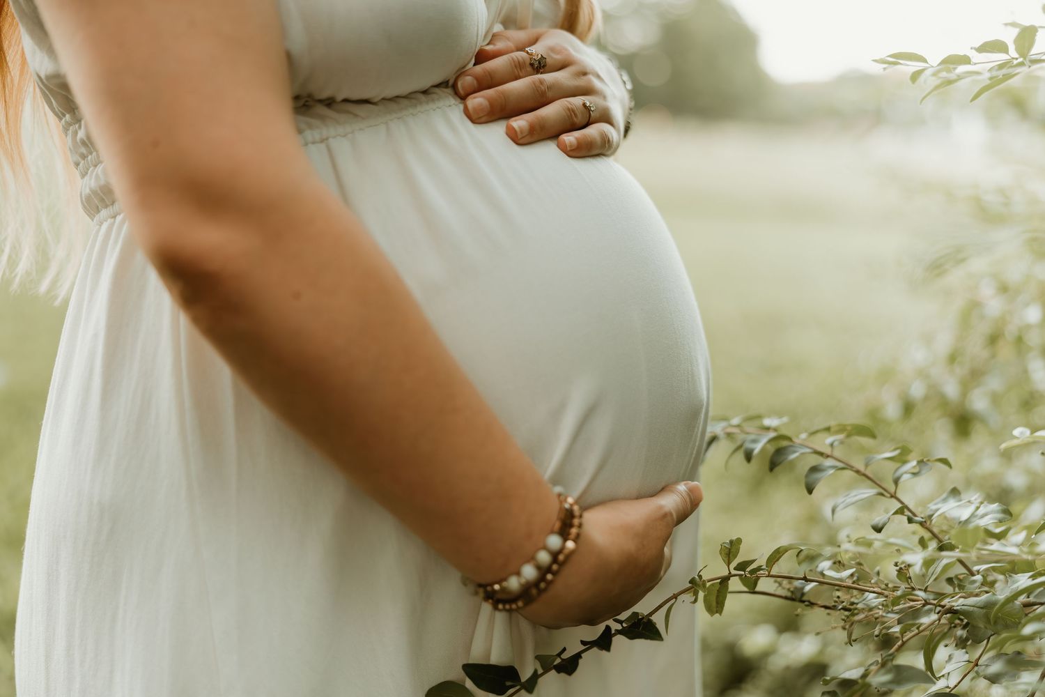 A pregnant woman in a white dress gently cradles her baby bump while standing in a sunlit meadow with wildflowers.
