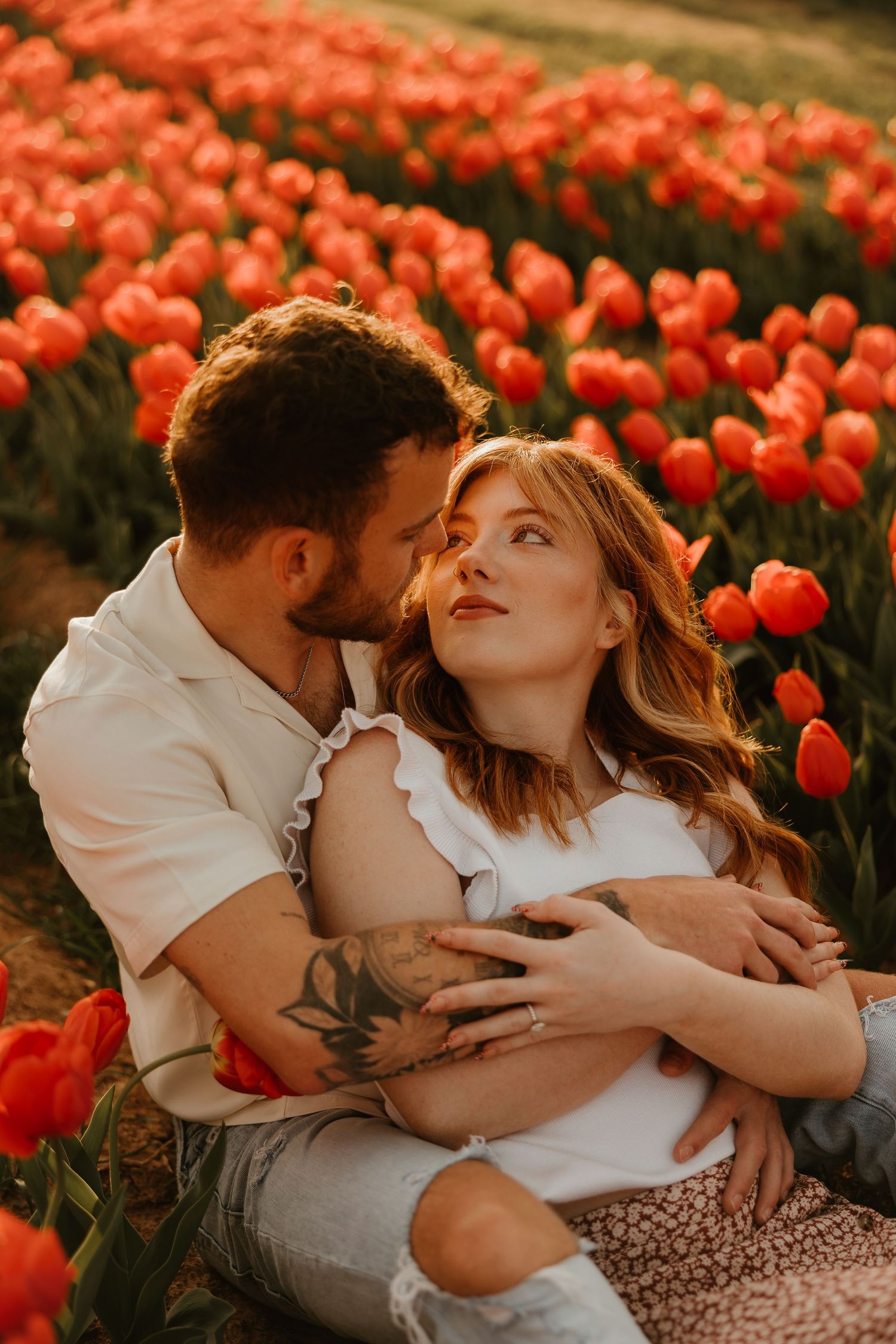 A romantic couple embraces in a vibrant red tulip field at sunset, dressed in white and creating an intimate moment.
