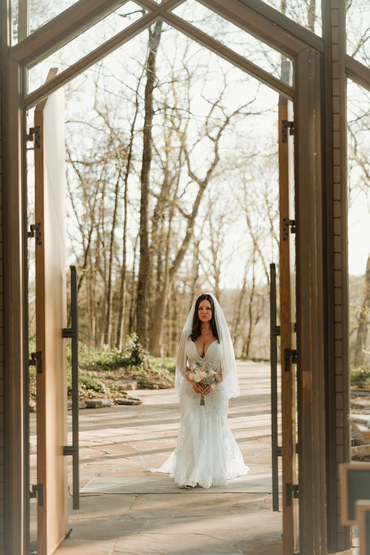 A bride in a white lace gown stands beneath a wooden archway framed by bare trees on a paved garden path.