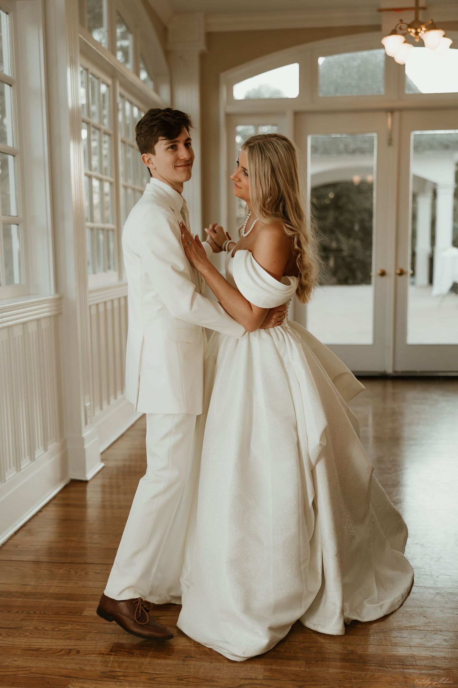 Couple in elegant white wedding attire dance together in sunlit room with hardwood floors and classic interior details.