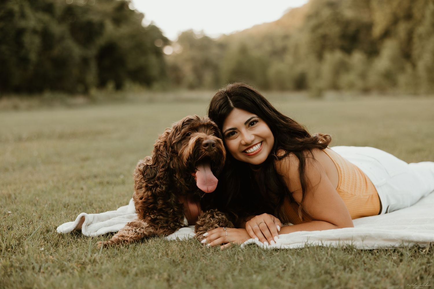 A woman in a yellow sweater lying on a blanket in a grassy field with a brown curly-haired dog at sunset.