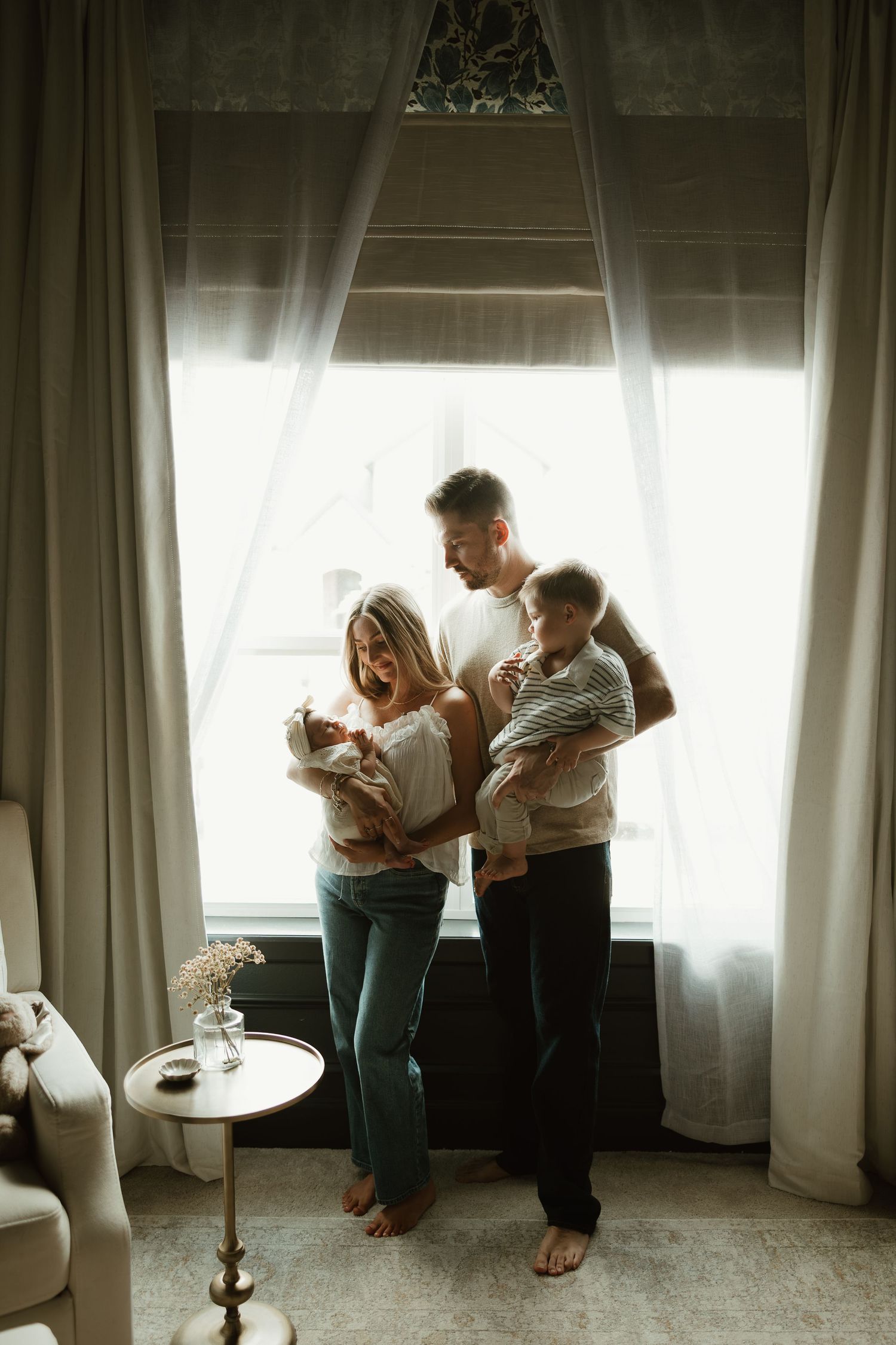 Young family with newborn and toddler standing by bright window in cozy living room, parents smiling lovingly.