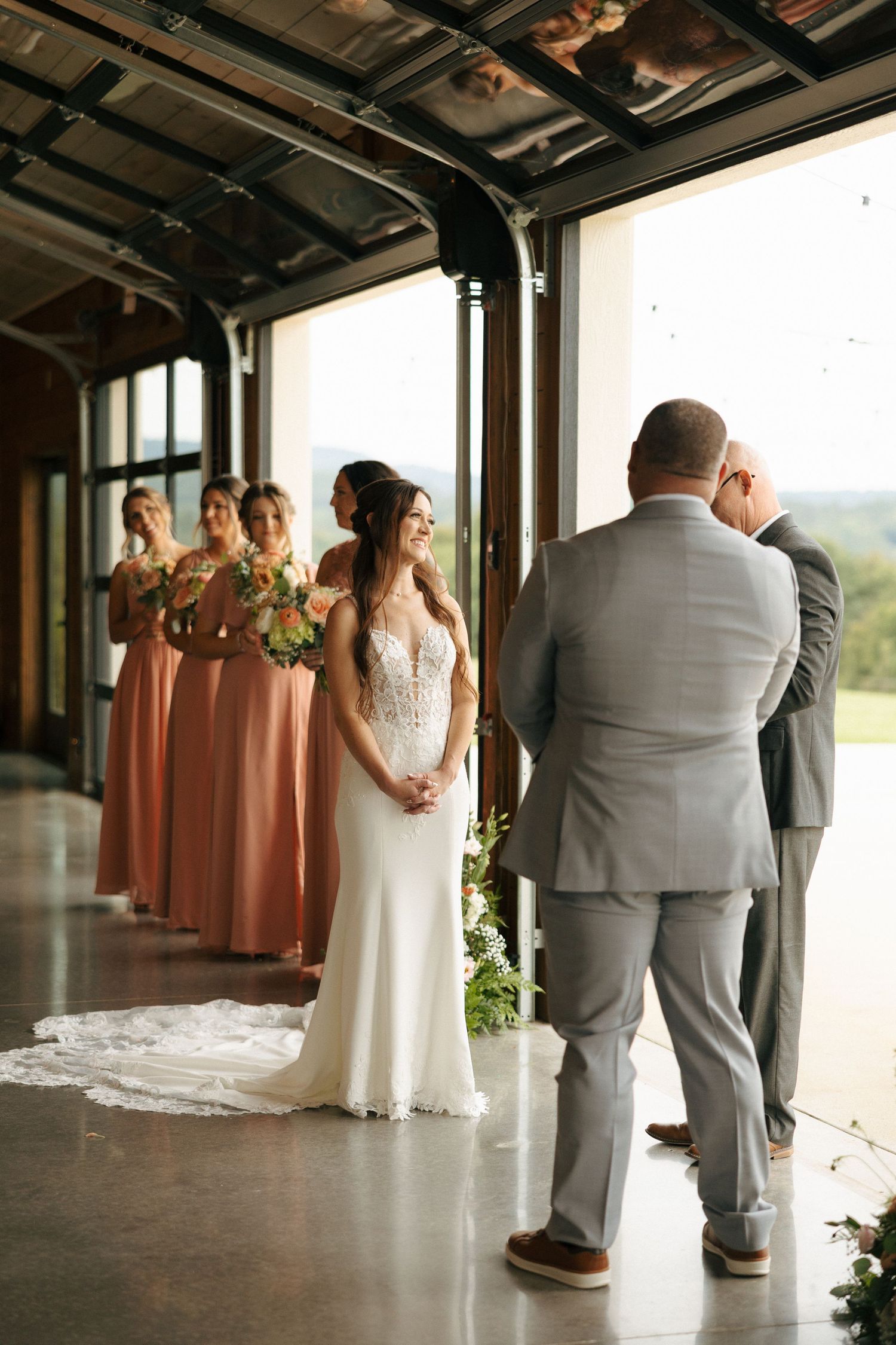 Wedding ceremony in an industrial venue with bridesmaids in coral dresses and a white bridal gown against large windows.