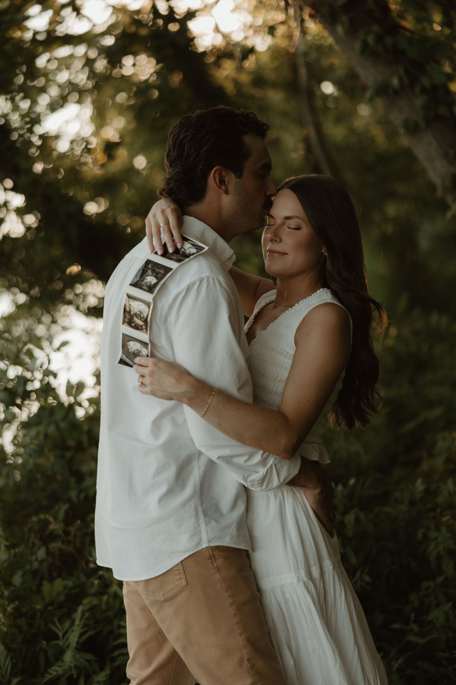 Couple embracing outdoors holding ultrasound photos, pregnancy announcement in a lush green garden setting.