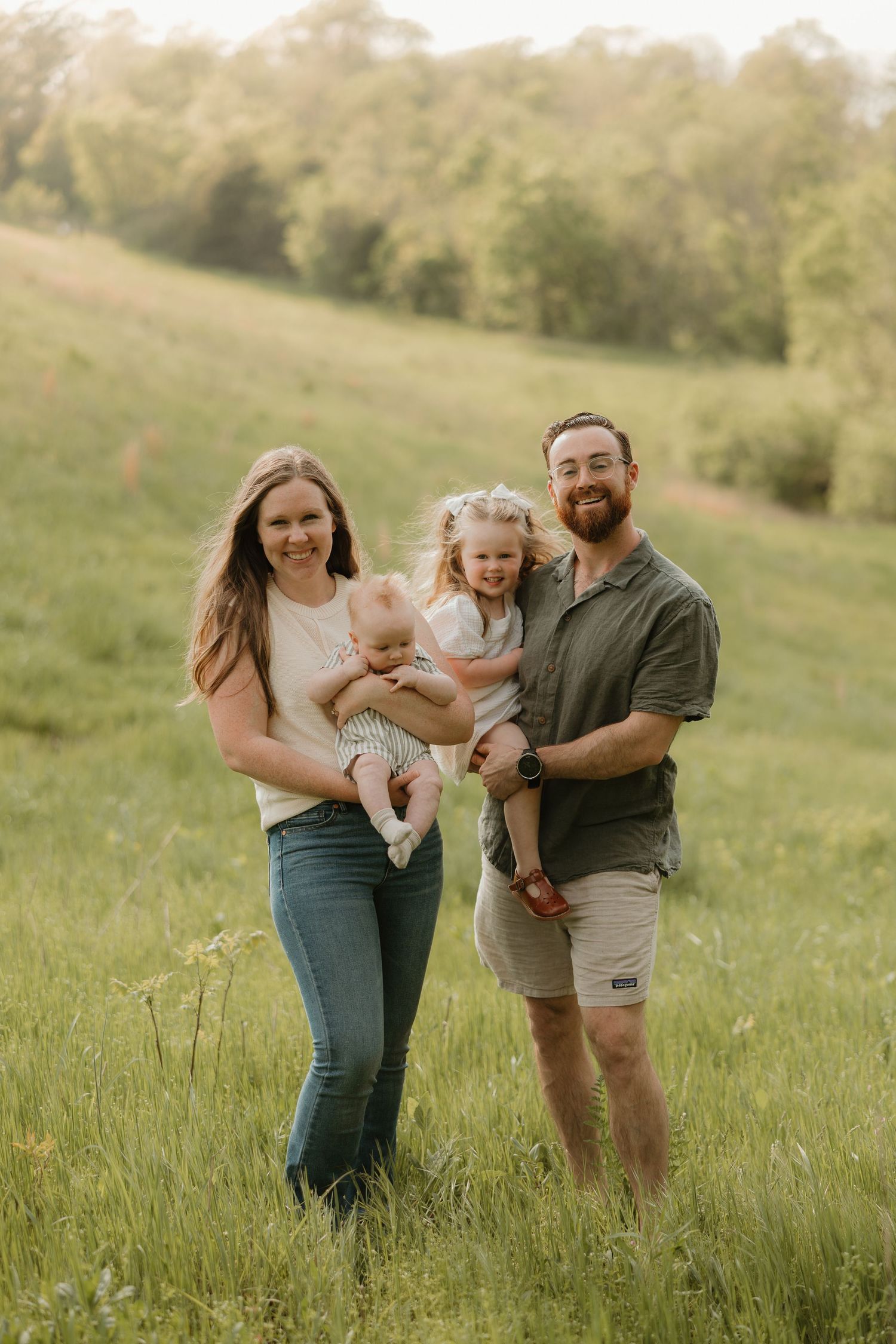 Family of three poses together in a sunny meadow during golden hour wearing casual clothing.